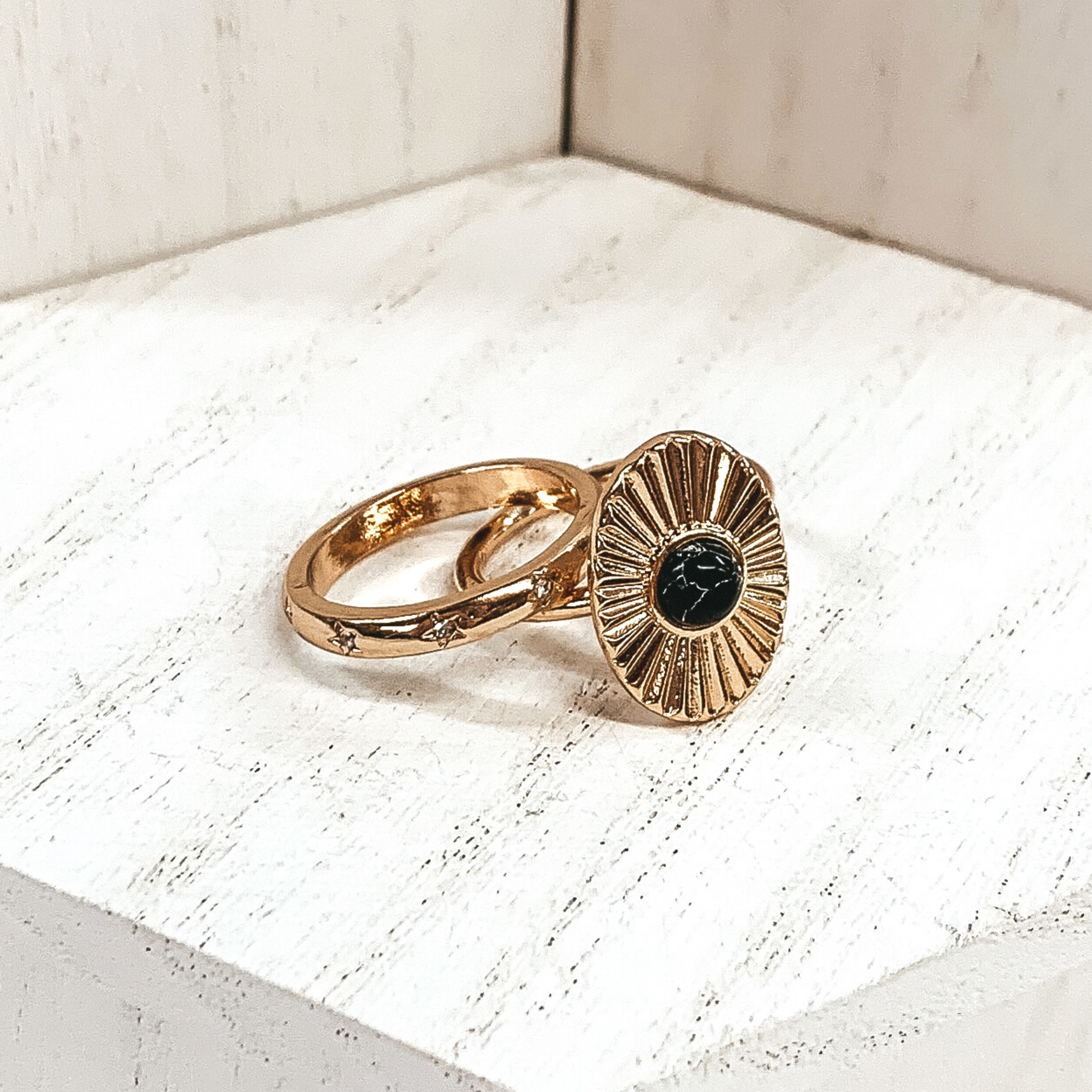 Both rings are gold in color. The first ring has an oval pendant with a starburst design on it and a center black stone. The second ring has tiny engraved stars with clear crystals in the center of them These rings are pictured on a white background.