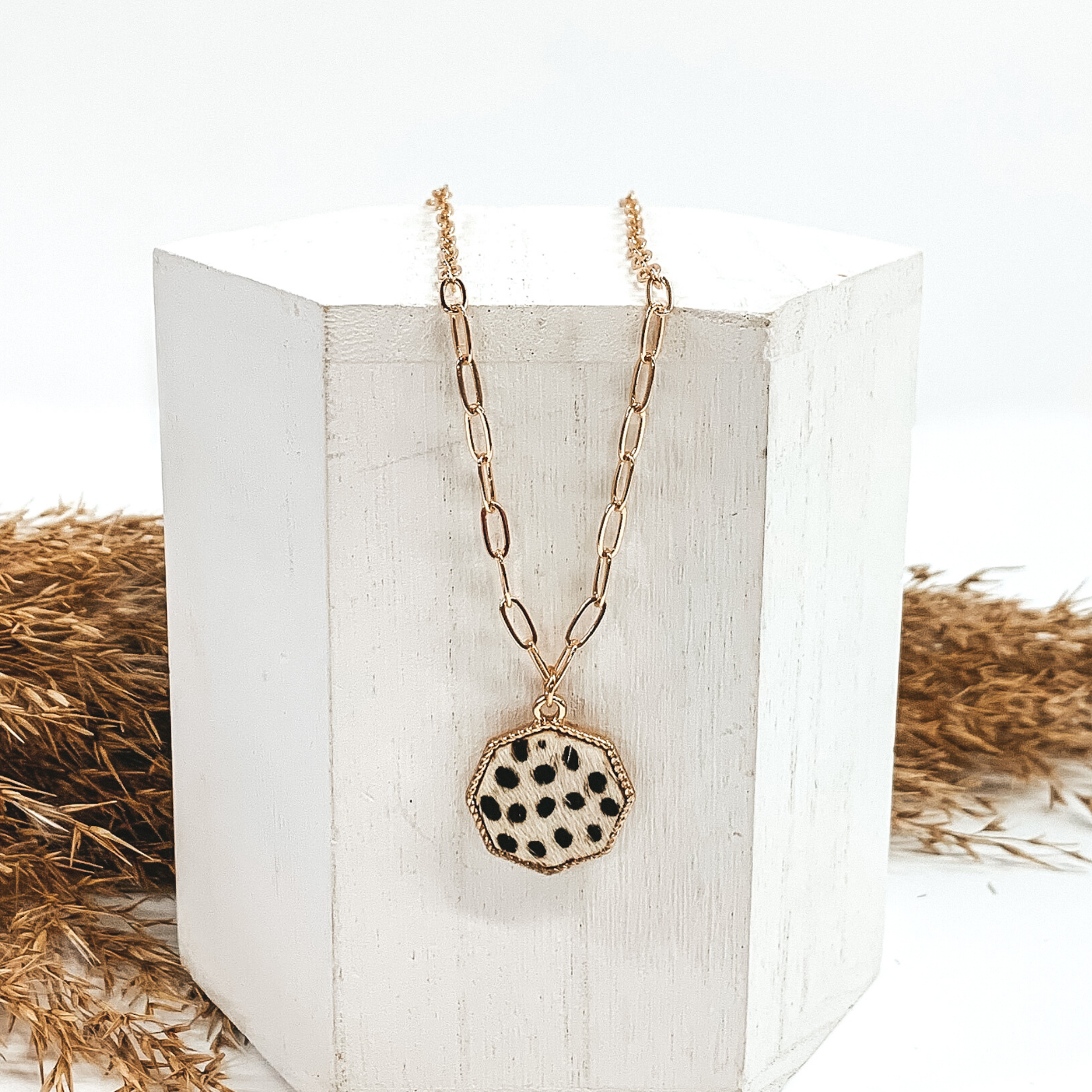 Gold paperclip chain with octagon pendant. The pendant has a white hide inlay with black dots. This necklace is pictured laying on a white block with tan floral behind it. This is all pictured on a white background. 