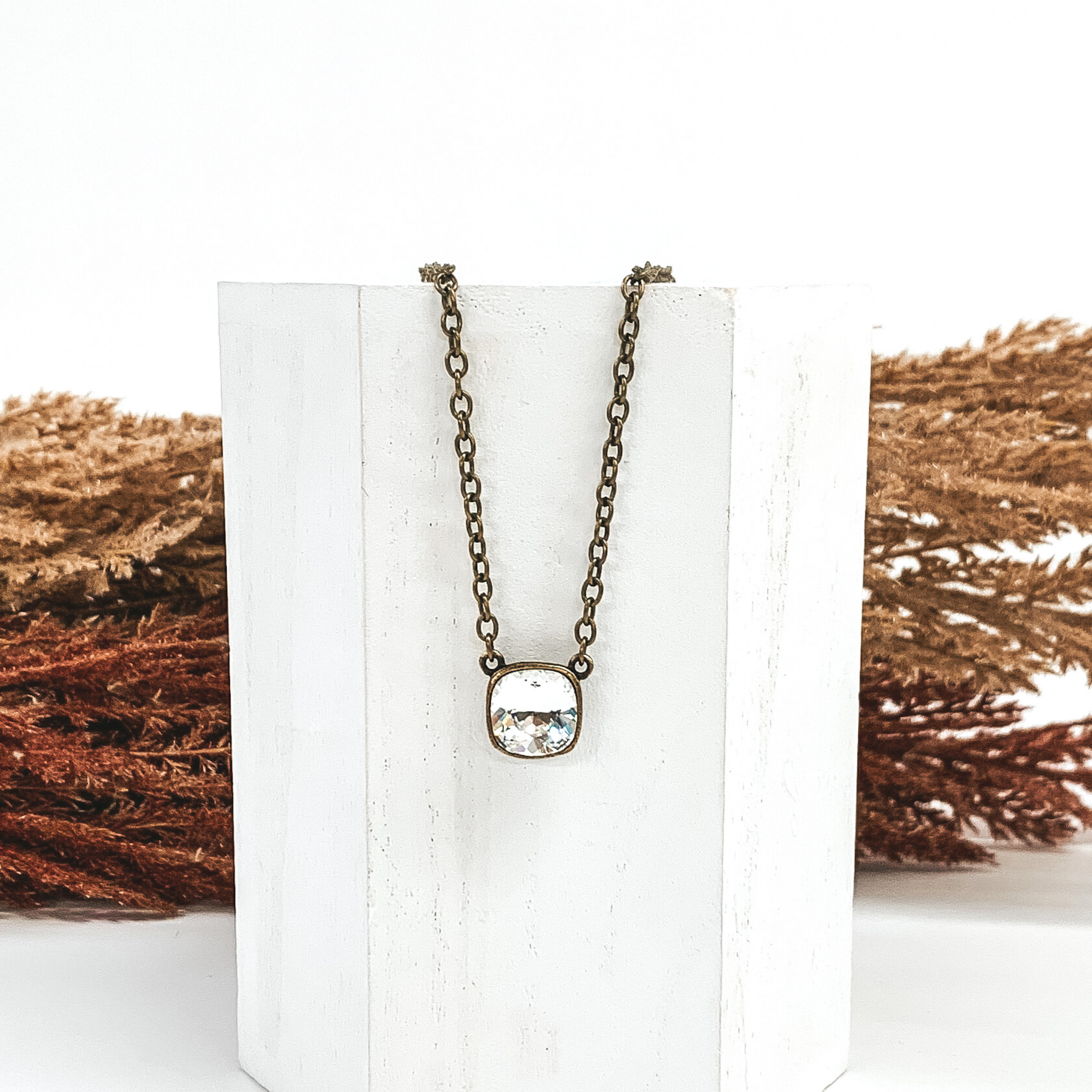 Bronzechained necklace with a square clear crystal pendant. The necklace is hanging on a white block in front of brown and tan decor