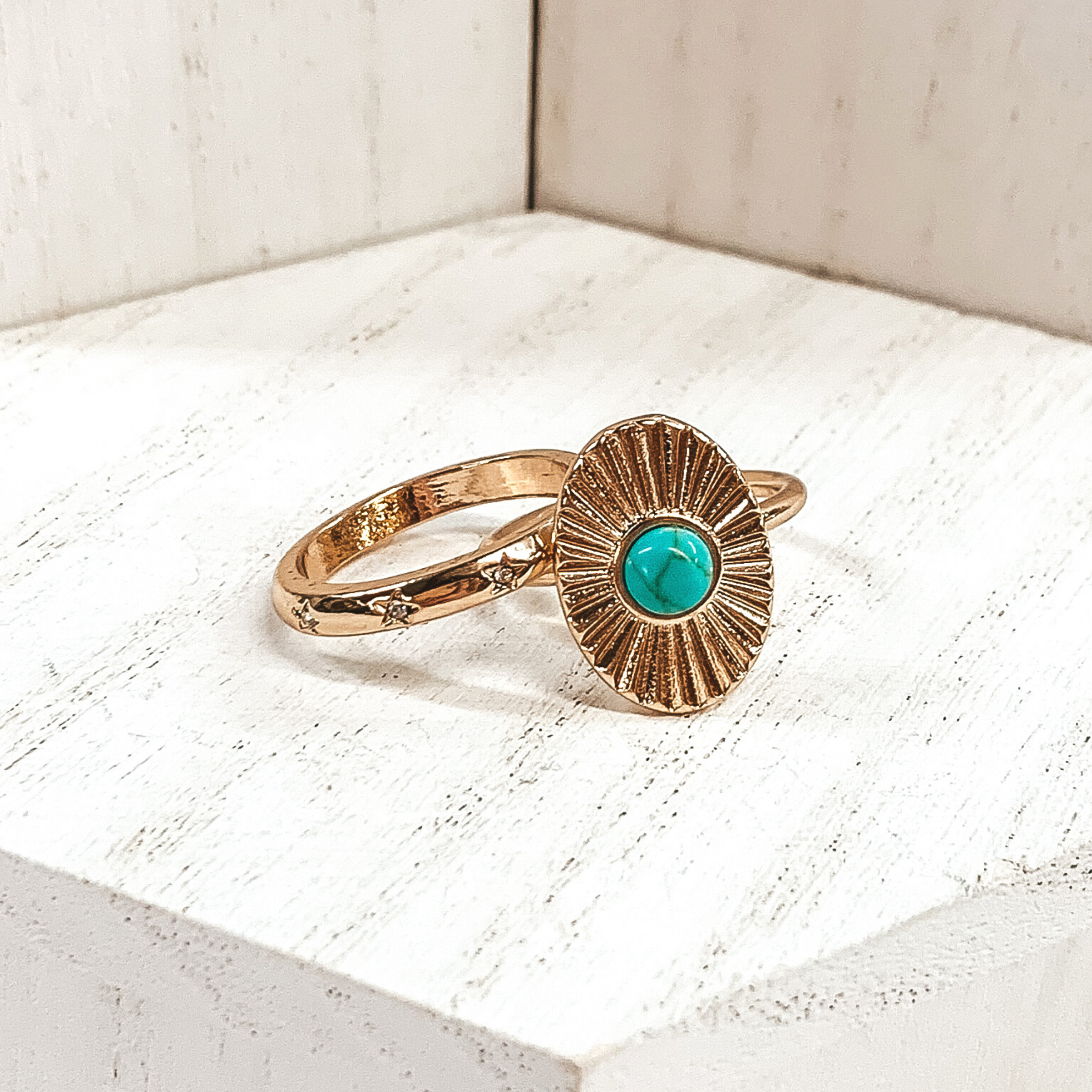 Both rings are gold in color. The first ring has an oval pendant with a starburst design on it and a center turquoise stone. The second ring has tiny engraved stars with clear crystals in the center of them These rings are pictured on a white background.