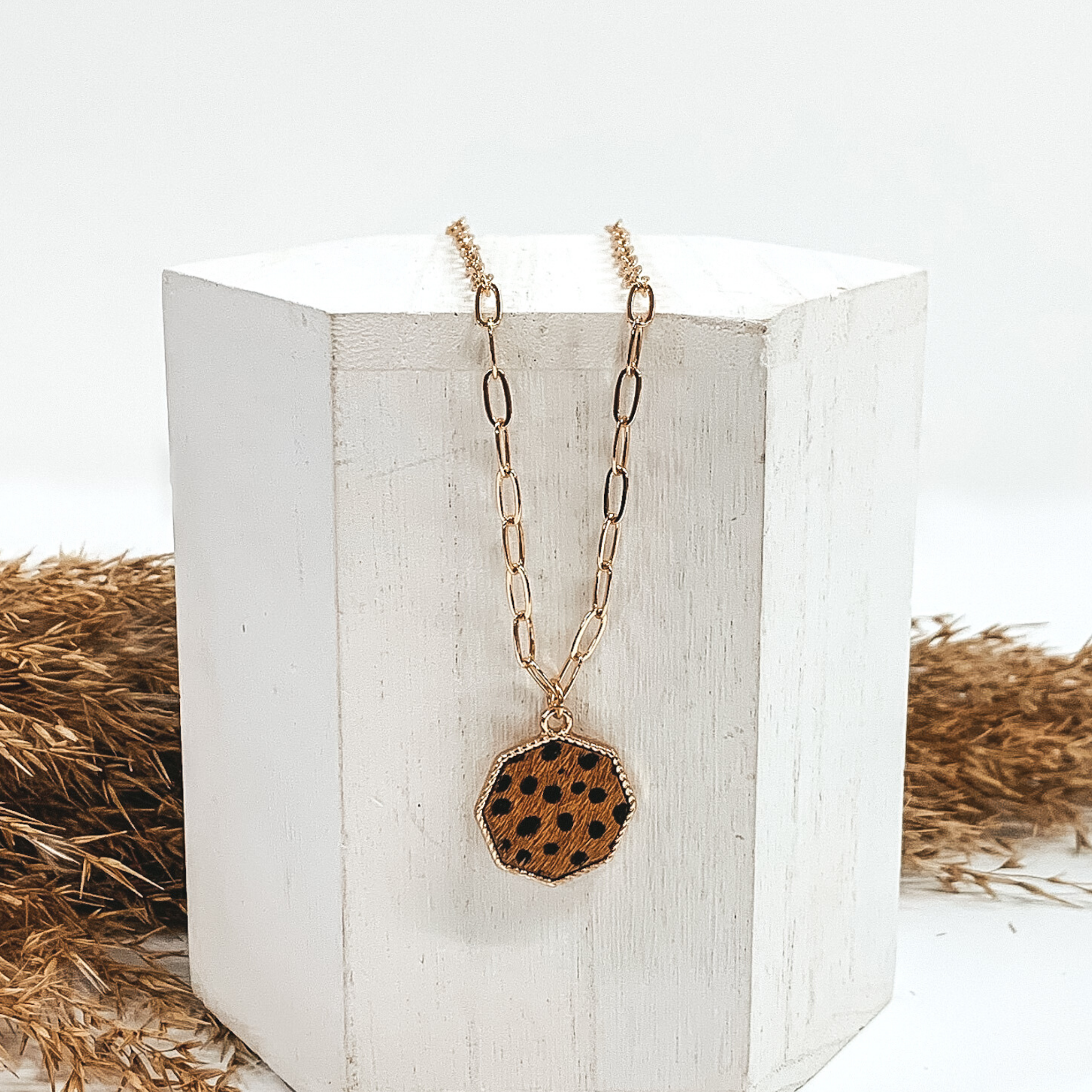 Gold paperclip chain with octagon pendant. The pendant has a brown hide inlay with black dots. This necklace is pictured laying on a white block with tan floral behind it. This is all pictured on a white background.