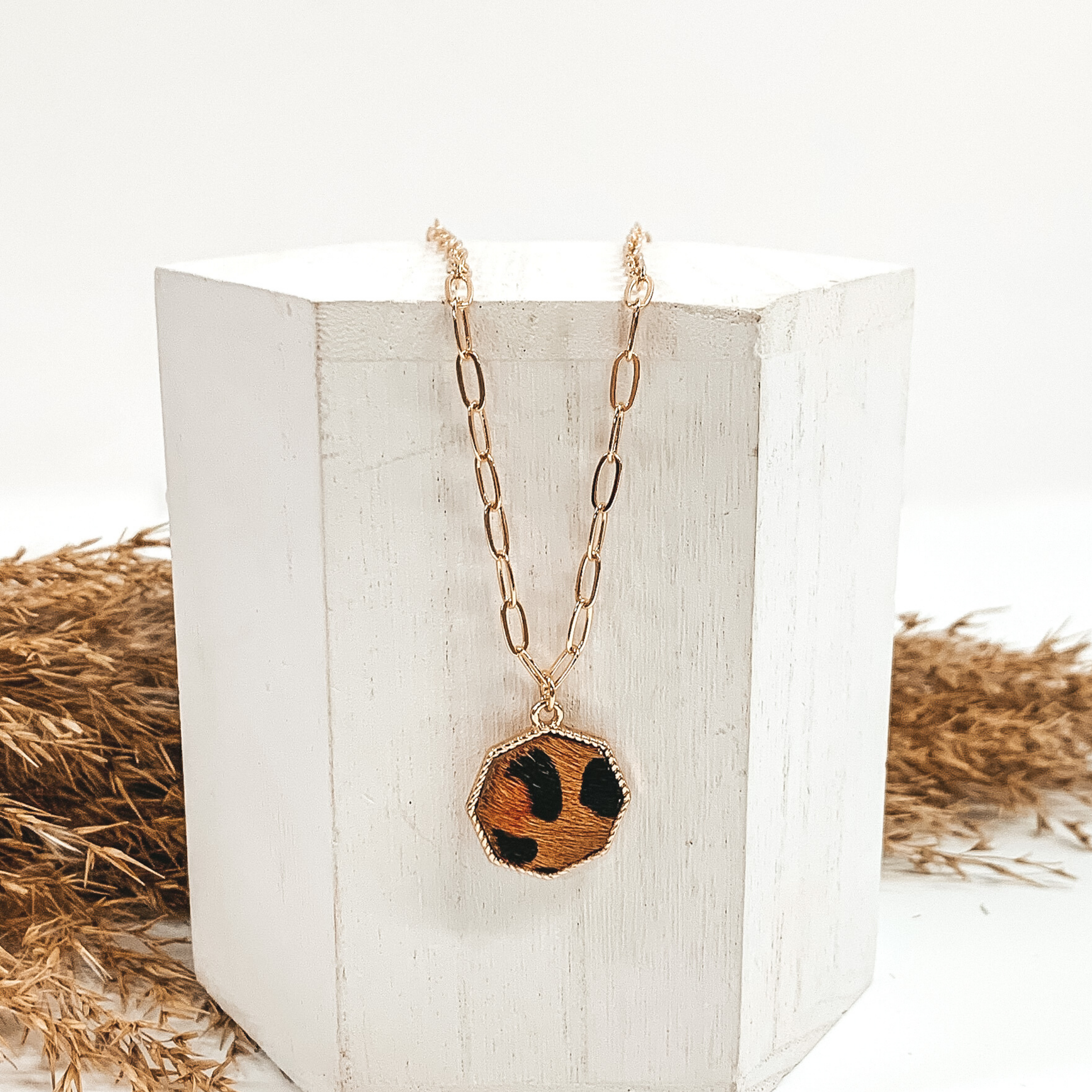Gold paperclip chain with octagon pendant. The pendant has a brown hide inlay with black dots. This necklace is pictured laying on a white block with tan floral behind it. This is all pictured on a white background.