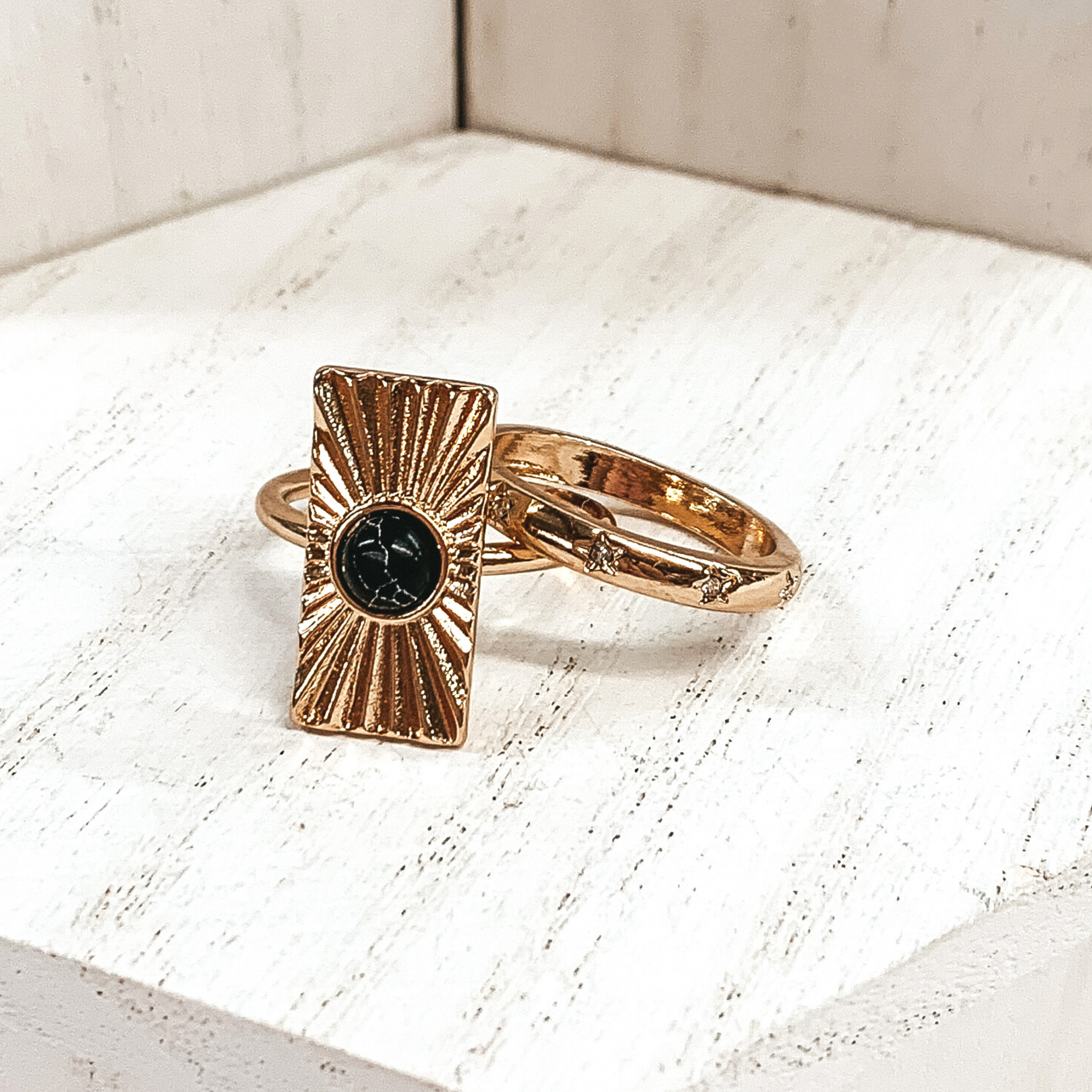 Both rings are gold in color. The first ring has an rectangle pendant with a starburst design on it and a center black stone. The second ring has tiny engraved stars with clear crystals in the center of them These rings are pictured on a white background.