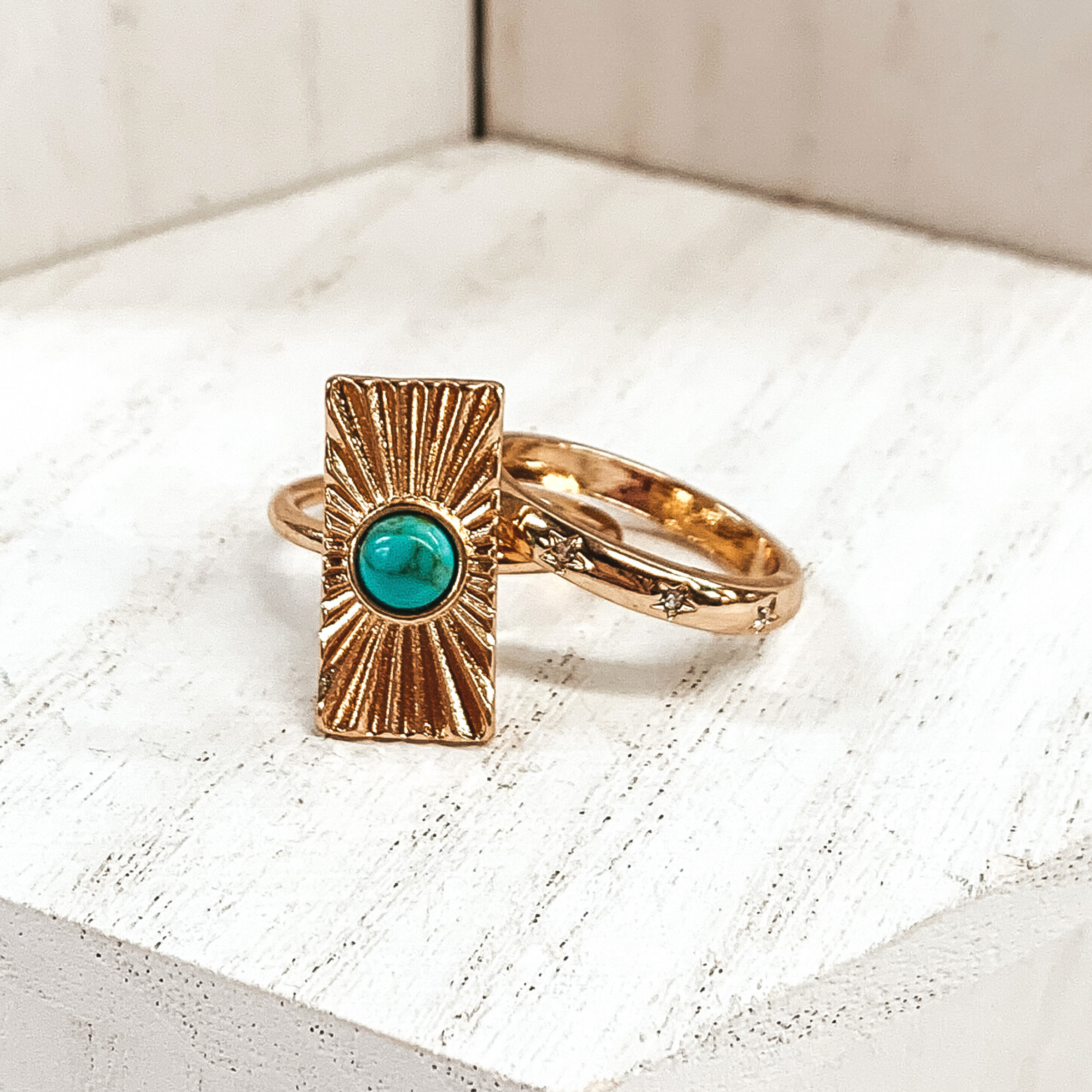 Both rings are gold in color. The first ring has an rectangle pendant with a starburst design on it and a center turquoise stone. The second ring has tiny engraved stars with clear crystals in the center of them These rings are pictured on a white background.