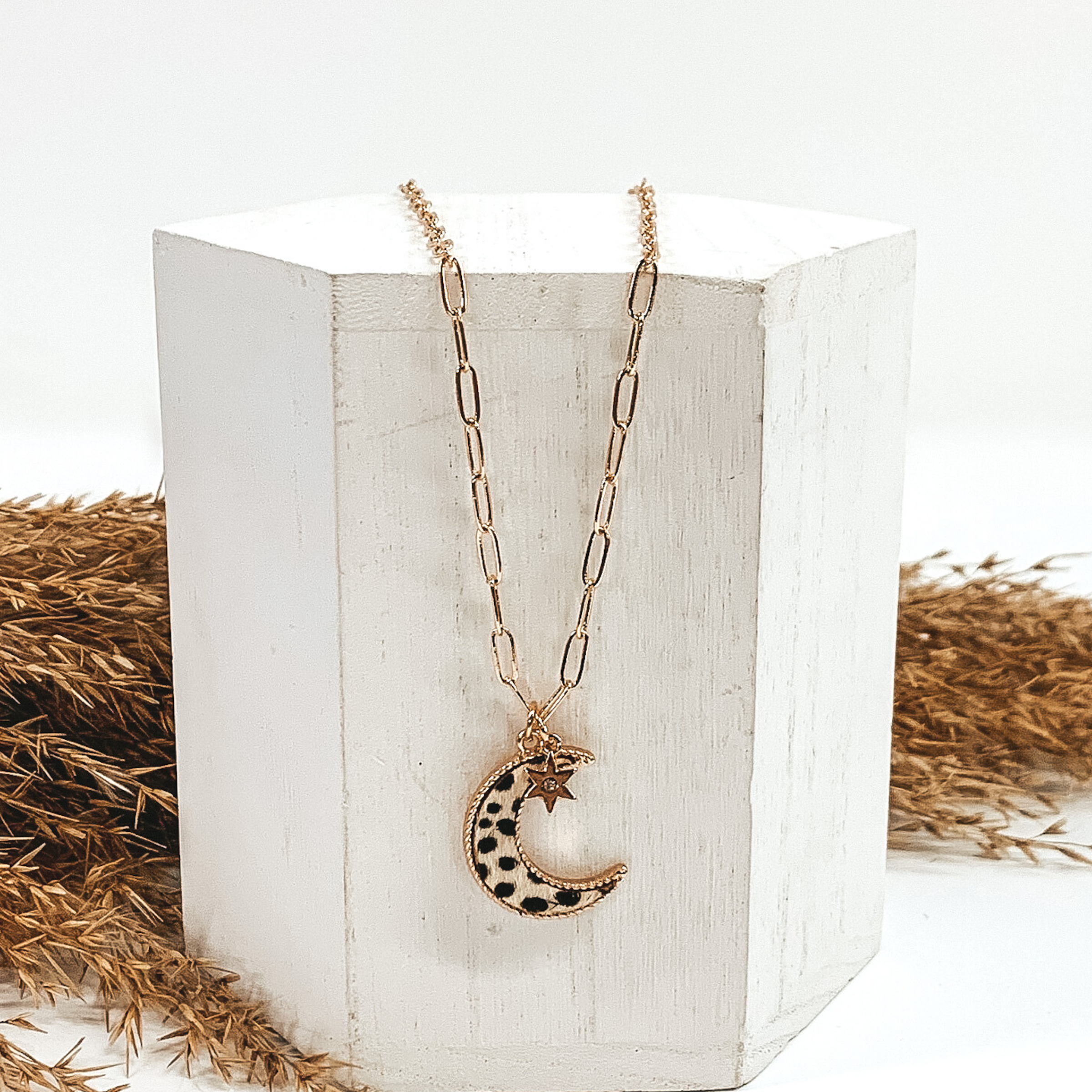 Gold paperclip chain with moon pendant and gold star charm with a tiny center crystal. The pendant has a white hide inlay with black dots. This necklace is pictured laying on a white block with tan floral behind it. This is all pictured on a white background.