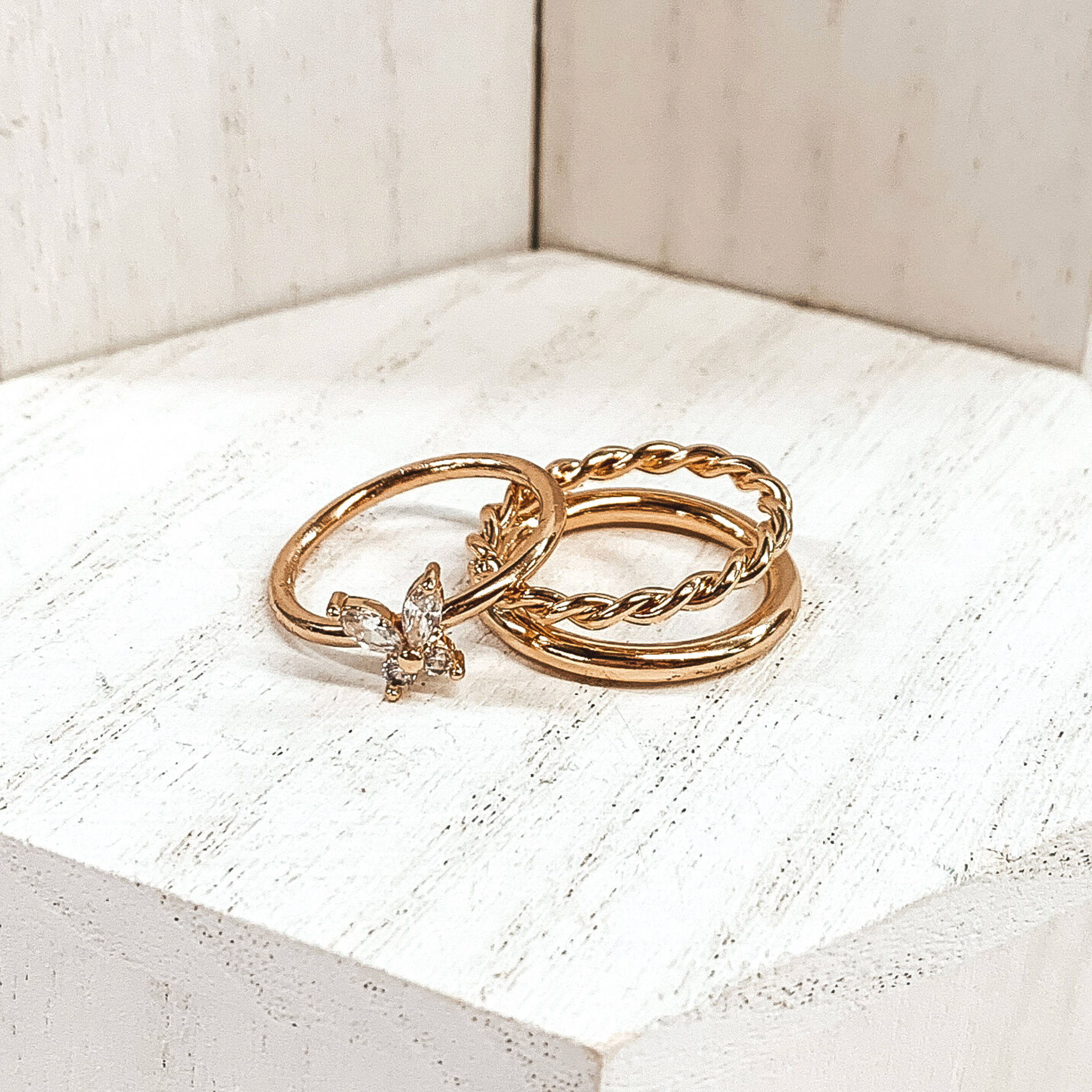 Both rings are gold in color. The double ring has one plain band and one twisted band. The ring that is leaning on it has a clear crystal butterfly pendant with a plain band. These rings are pictured on a white background.