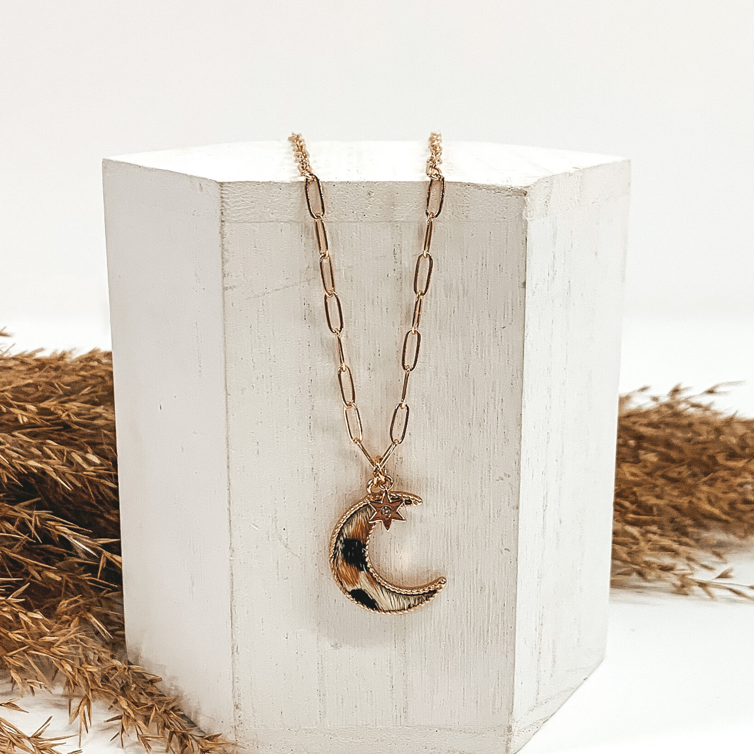 Gold paperclip chain with octagon pendant. The pendant has a white hide animal print inlay. This necklace is pictured laying on a white block with tan floral behind it. This is all pictured on a white background.