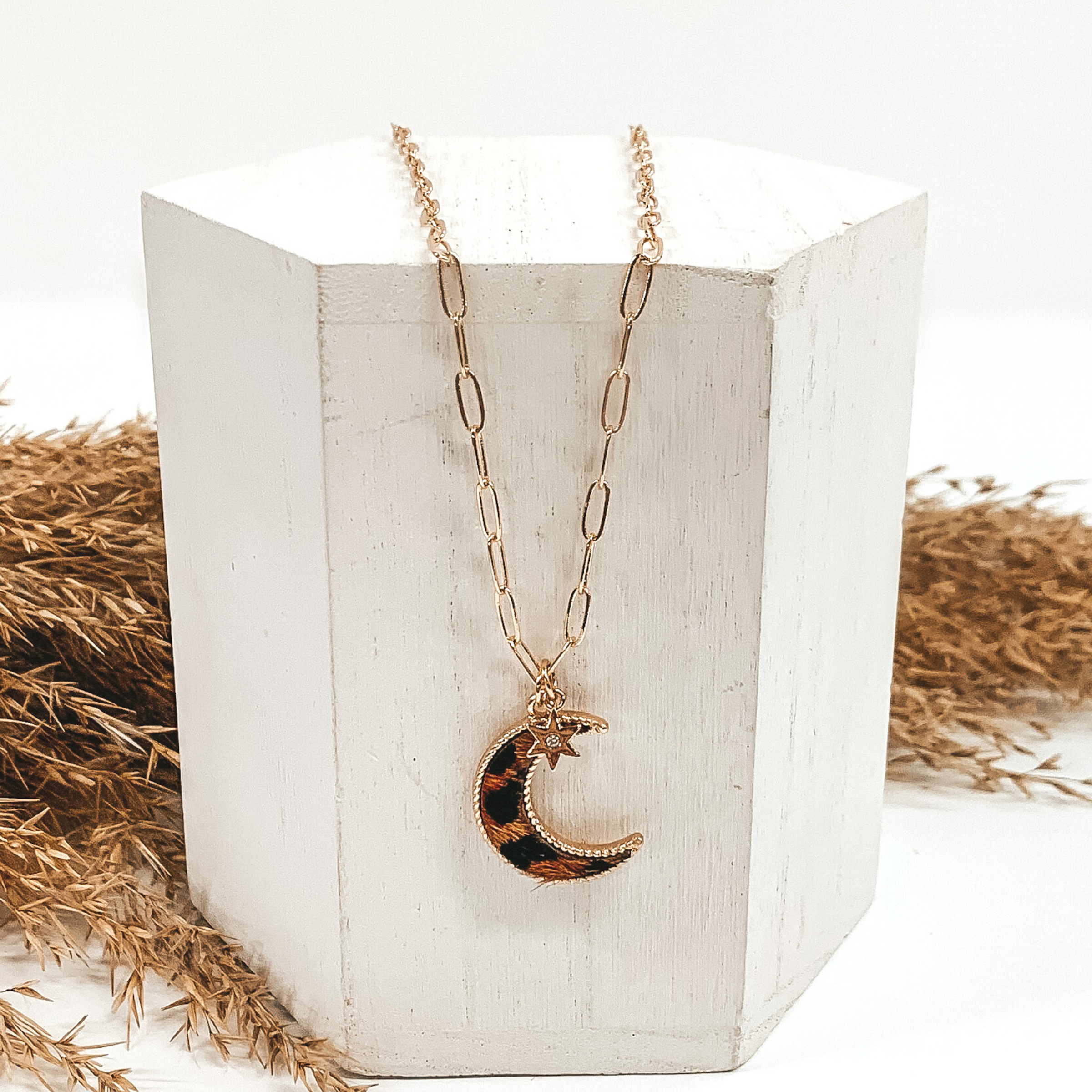 Gold paperclip chain with octagon pendant. The pendant has a brown hide animal print inlay. This necklace is pictured laying on a white block with tan floral behind it. This is all pictured on a white background.