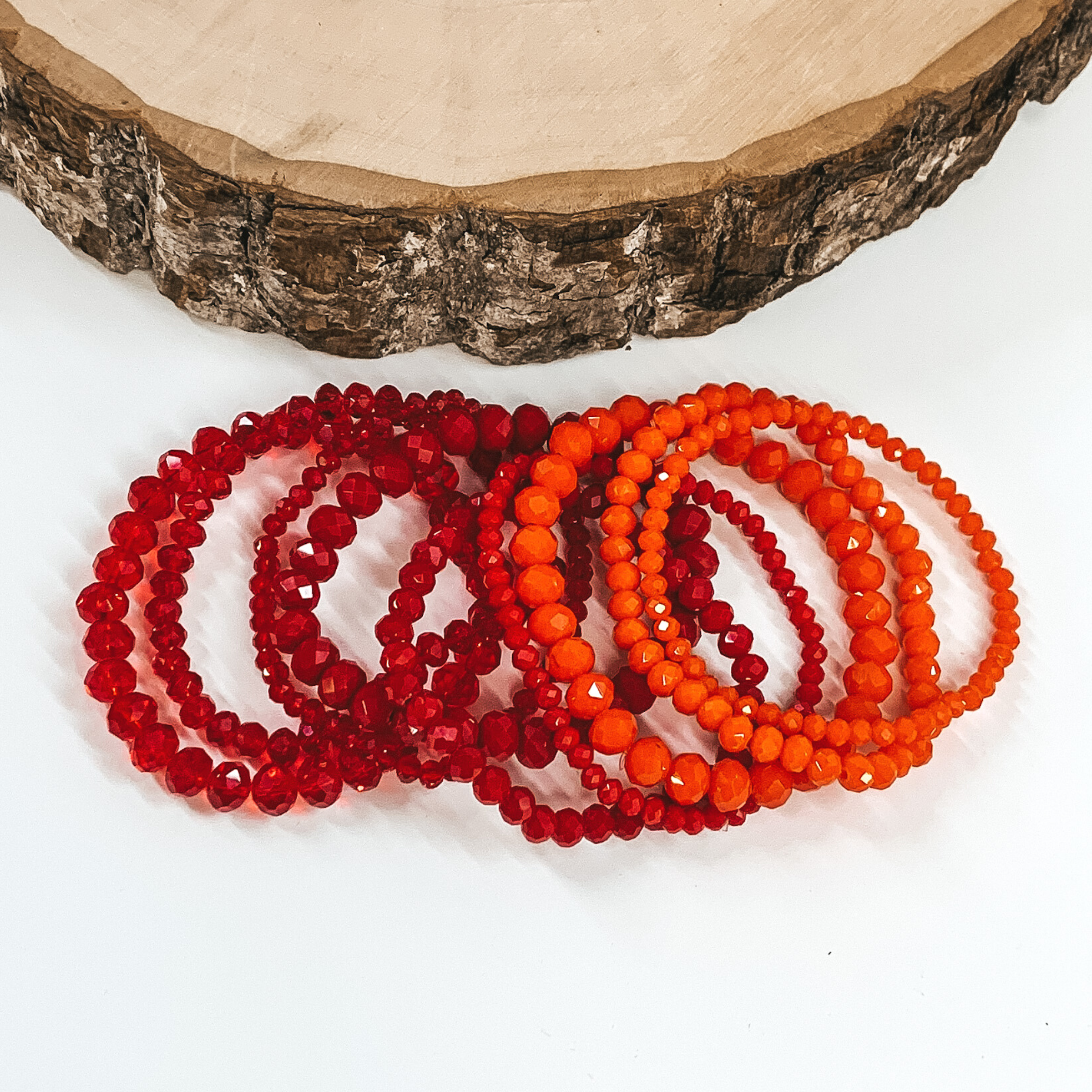 Set of nine crystal beaded bracelets. You have three shiny red, three matte red, and three matte orange. Each color has three different size beaded bracelets. these bracelets are laying on top of each other in a line on a white background with a piece of wood at the top of the picture. 