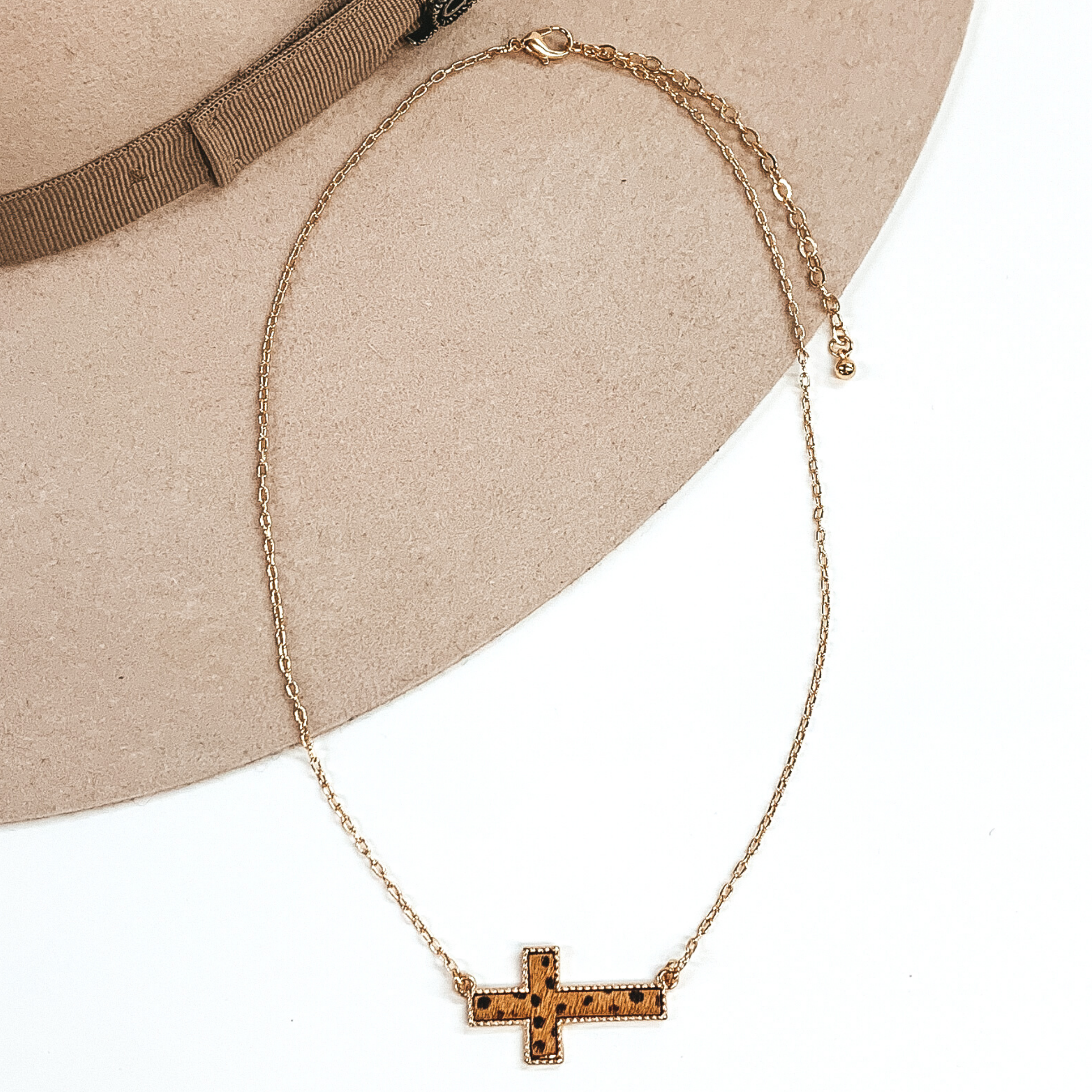 Gold paperclip chain with a cross pendant. The pendant has a brown hide inlay with black dots. this necklace is pictured on a white and beige background.