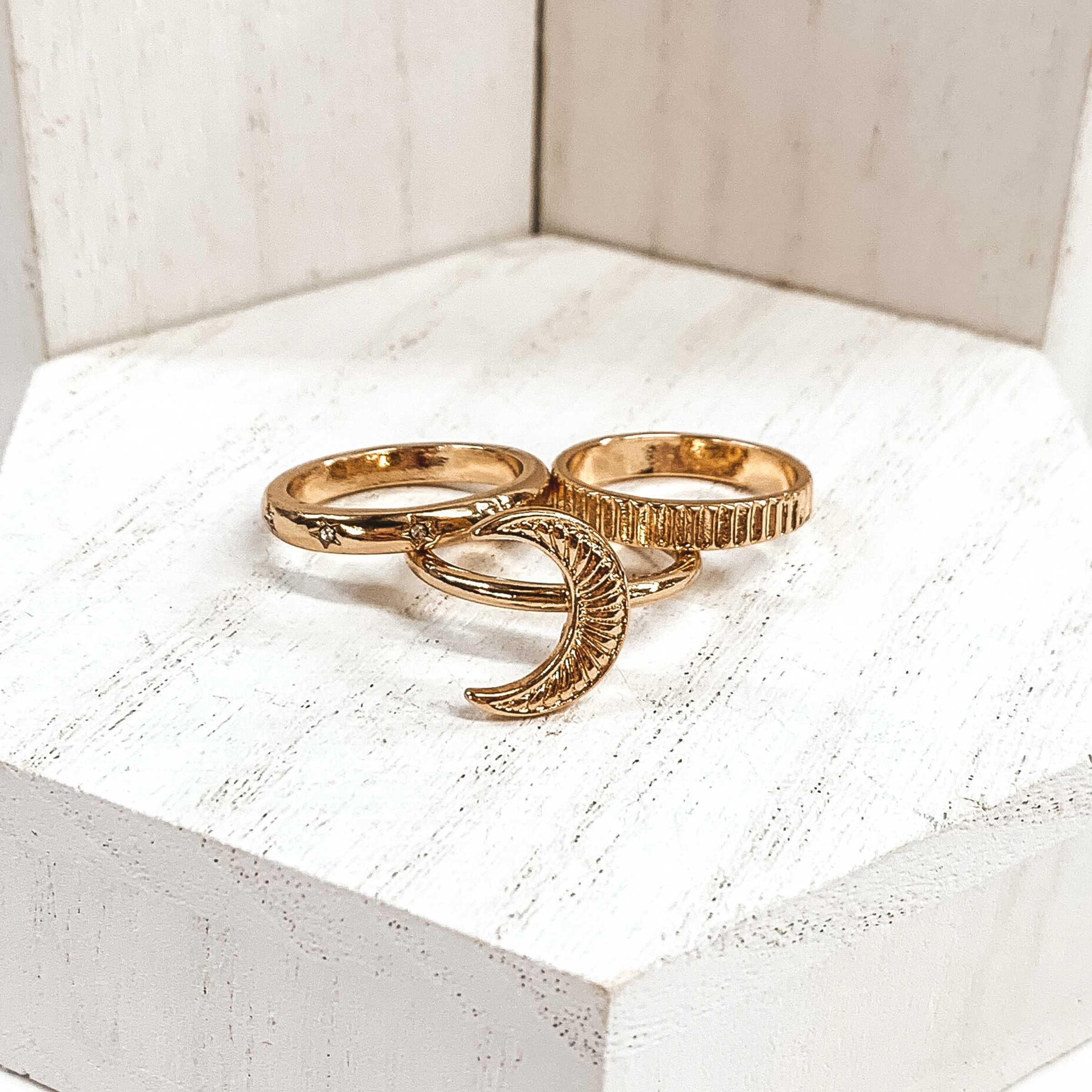 Set of three gold rings. One has a moon pendant, one is an indented striped ring, and the last on has tiny engraved star with tiny clear crystals in the middle. These rings are pictured on a white background.