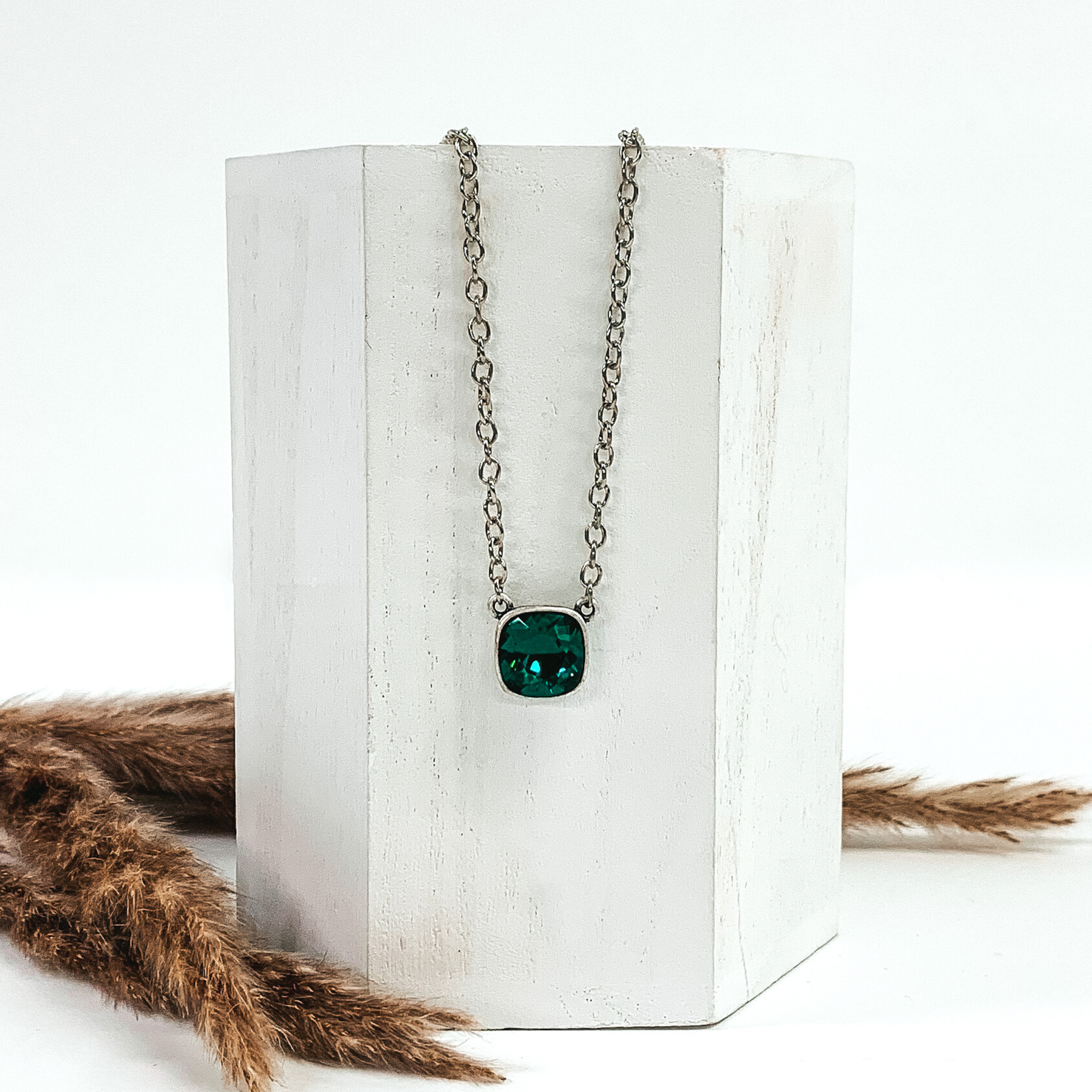 Silver chained necklace with a square emerald crystal. The necklace is hanging on a white block next the brown floral decor