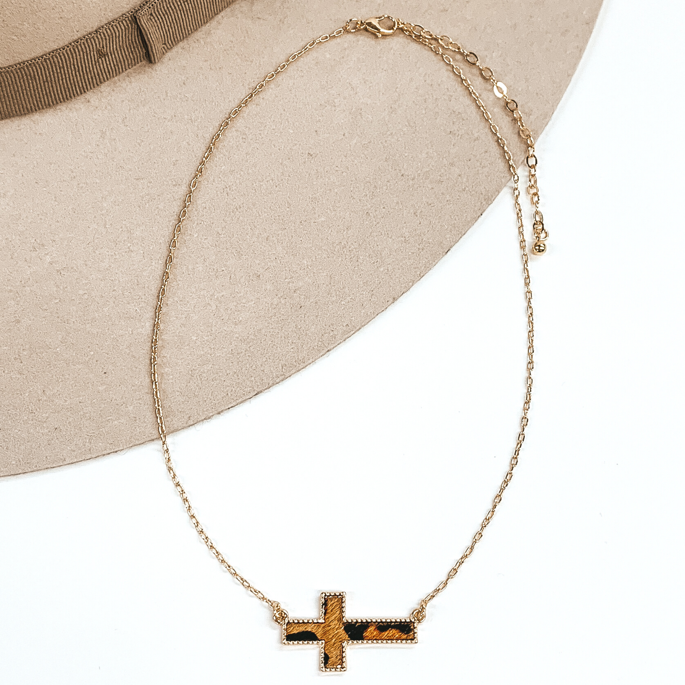 Gold paperclip chain with a cross pendant. The pendant has a brown hide inlay with an animal print. This necklace is pictured on a white and beige background.