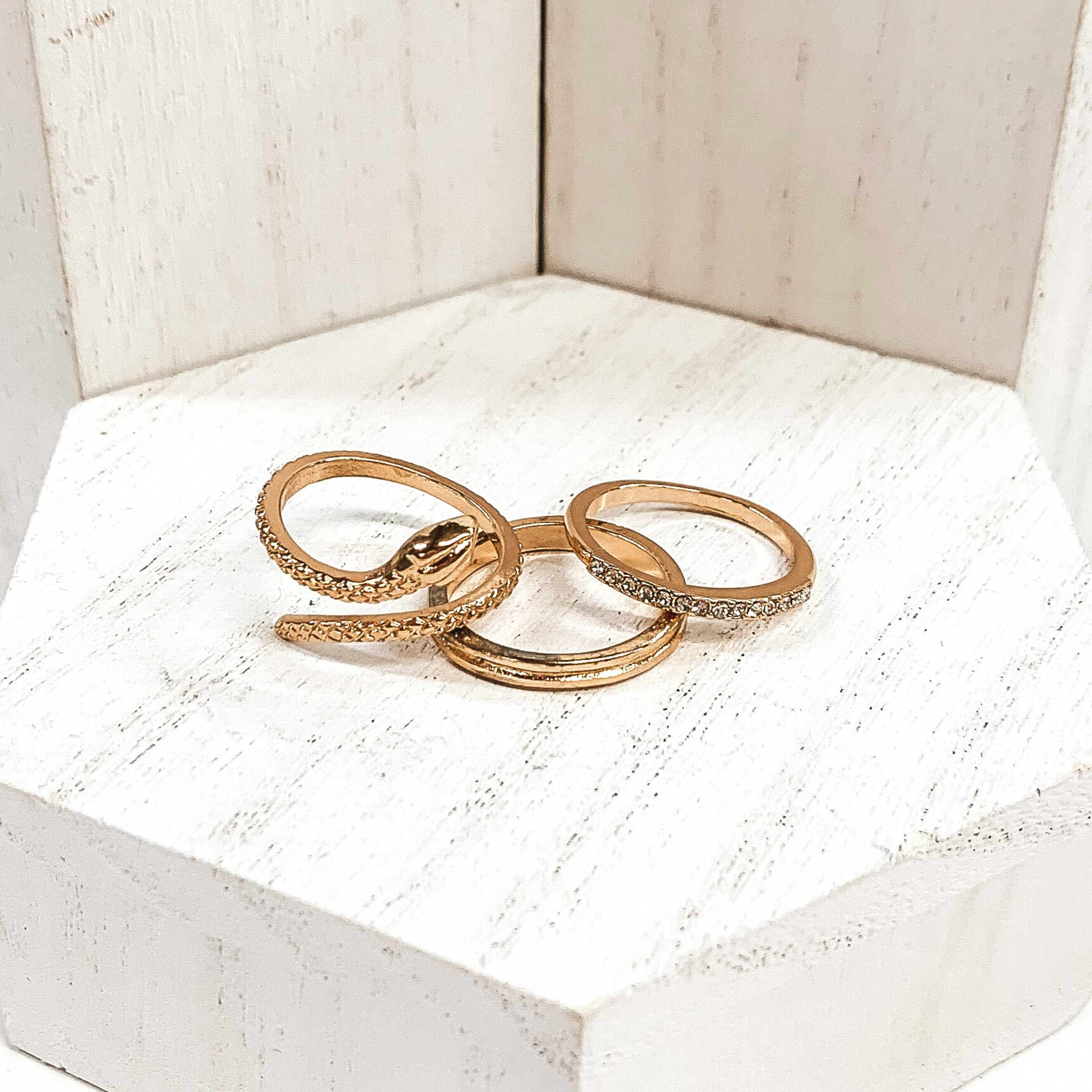 This gold ring set is very edgy. It includes one snake ring, one double layered ring, and one ring with a row of crystals. These rings are pictured on a white background.