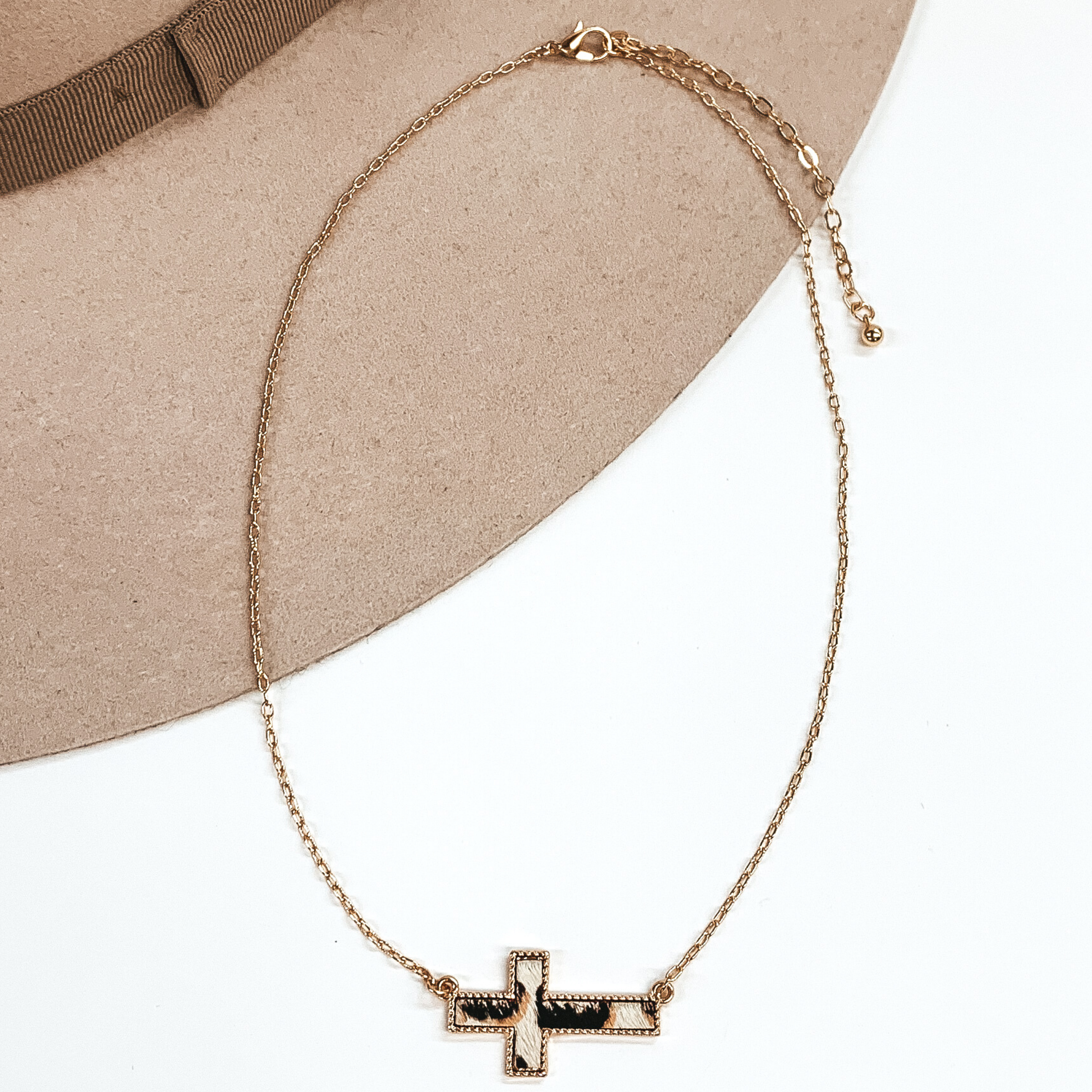 Gold paperclip chain with a cross pendant. The pendant has a white hide inlay with an animal print. This necklace is pictured on a white and beige background.
