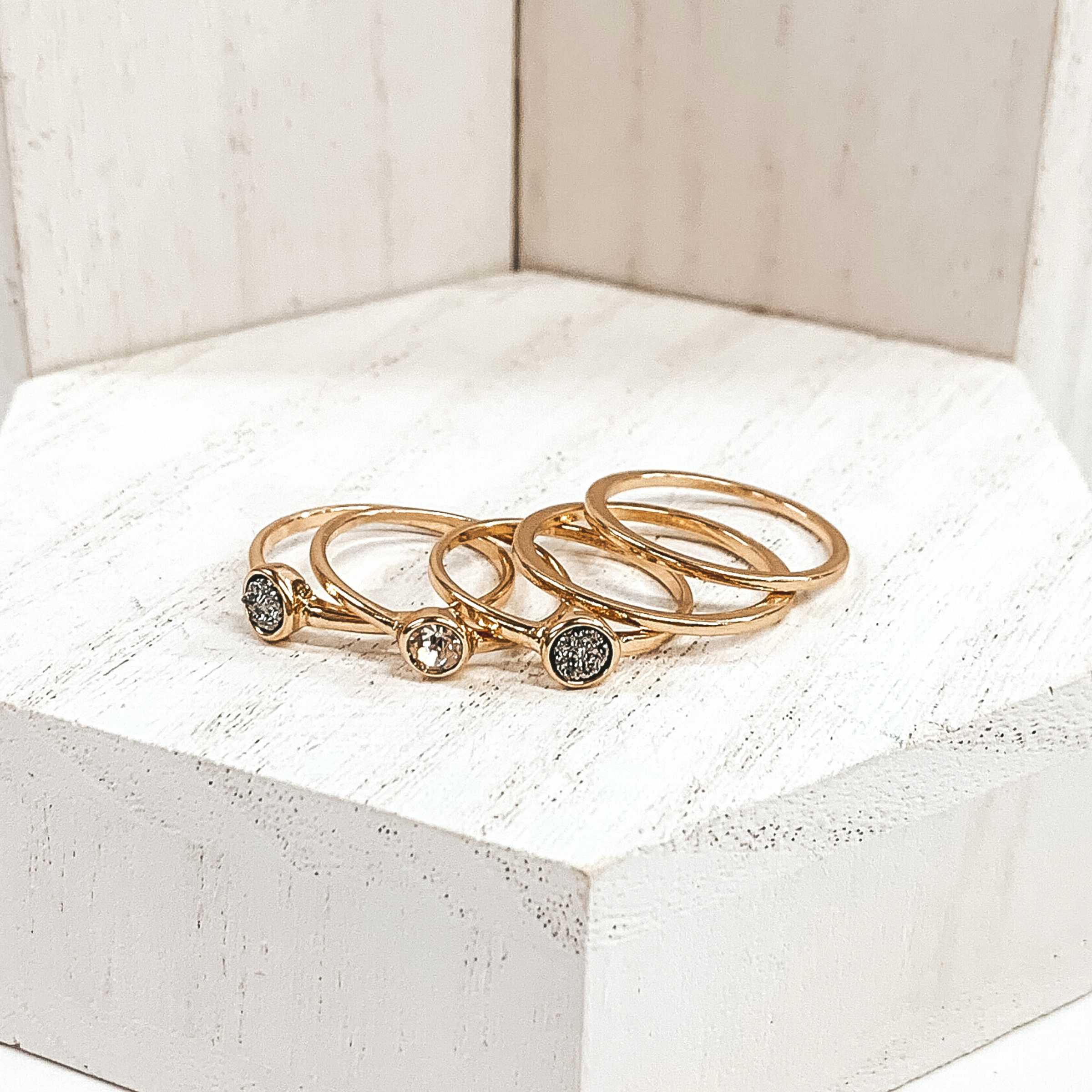 This is a set of 5 gold rings. Two rings are plain gold bands, one has a clear crystal stone, and the other two rings have grey druzy stones. These rings are pictured on a white background.