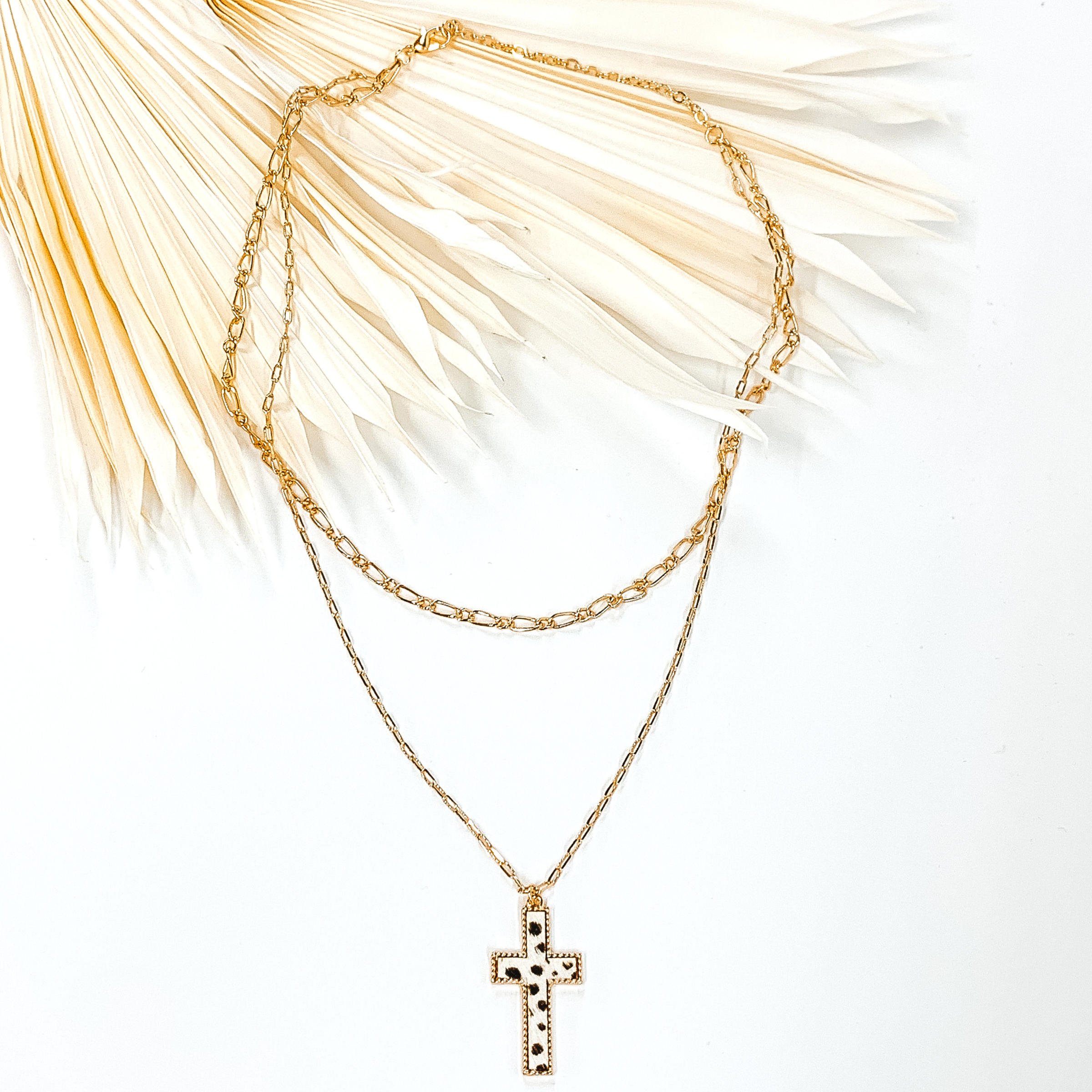Gold paperclip doubled chain with a cross pendant. The pendant has a white hide inlay with a dotted print. This necklace is pictured laying on cream leaf on a white background.
