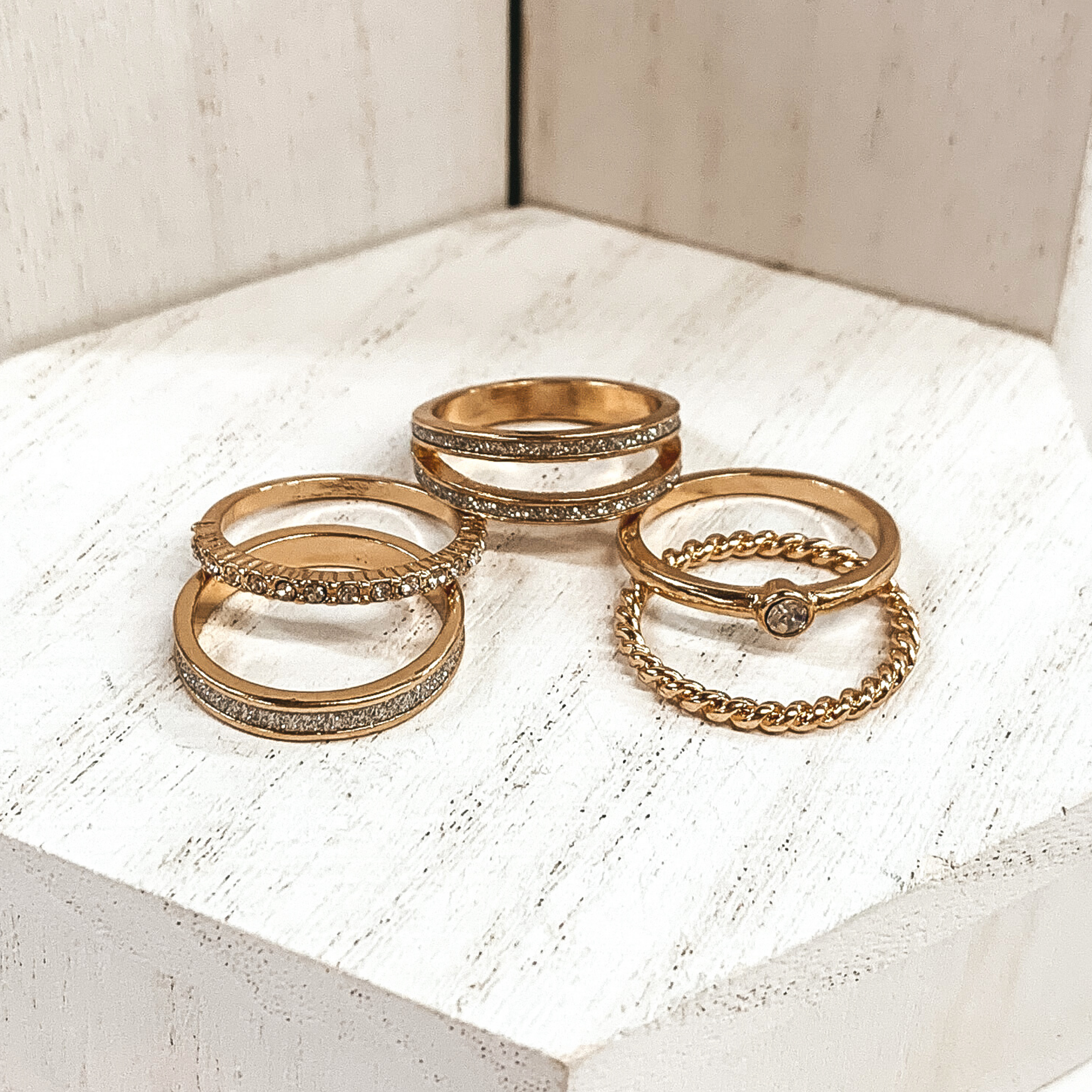 This is a set of five gold rings. All of them but one ring has crystals in different varieties on them. The single ring that does not have crystals is a twisted ring. These rings are pictured on a white background.