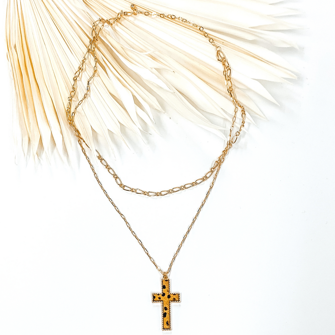 Gold paperclip doubled chain with a cross pendant. The pendant has a brown hide inlay with a dotted print. This necklace is pictured laying on cream leaf on a white background.