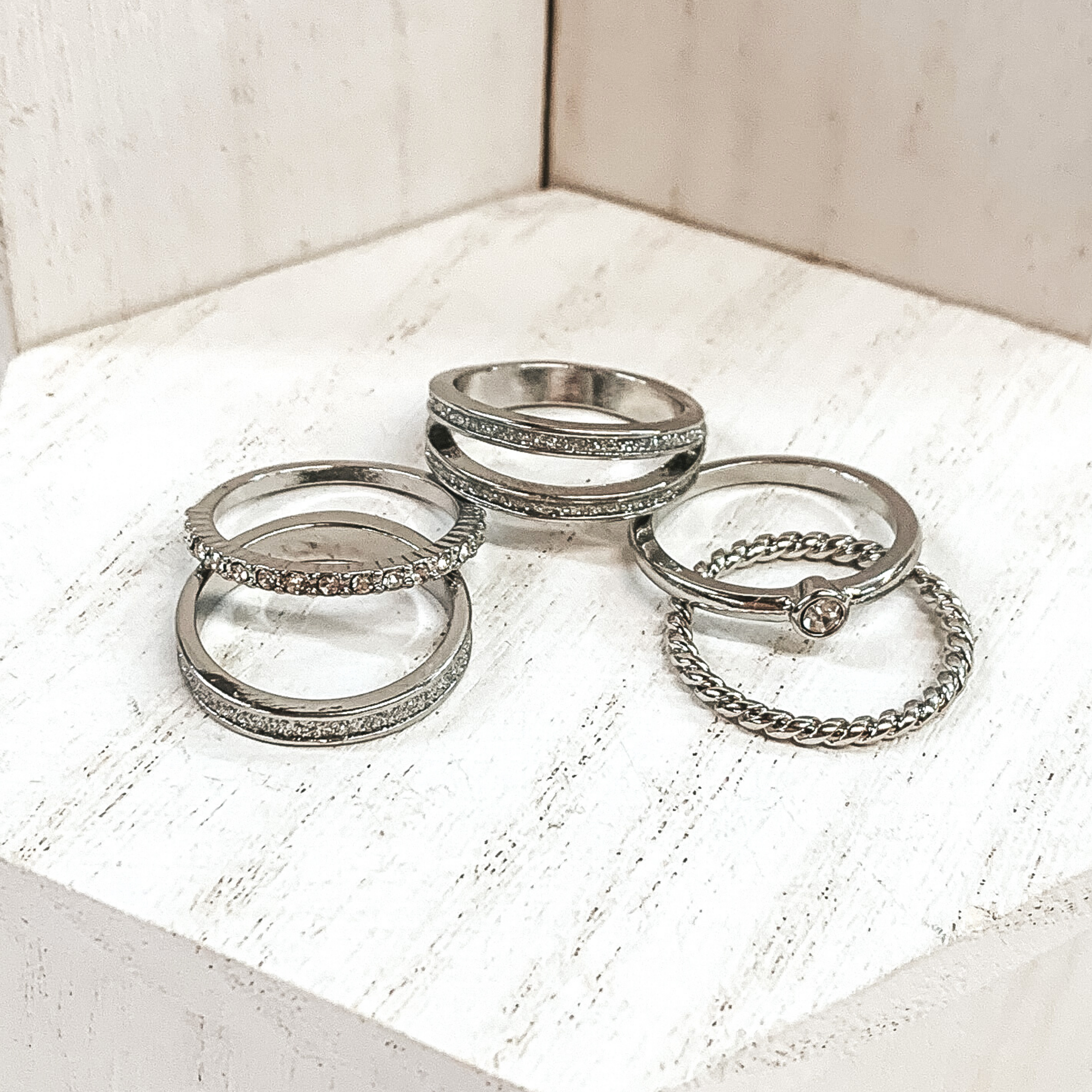 This is a set of five silver rings. All of them but one ring has crystals in different varieties on them. The single ring that does not have crystals is a twisted ring. These rings are pictured on a white background.