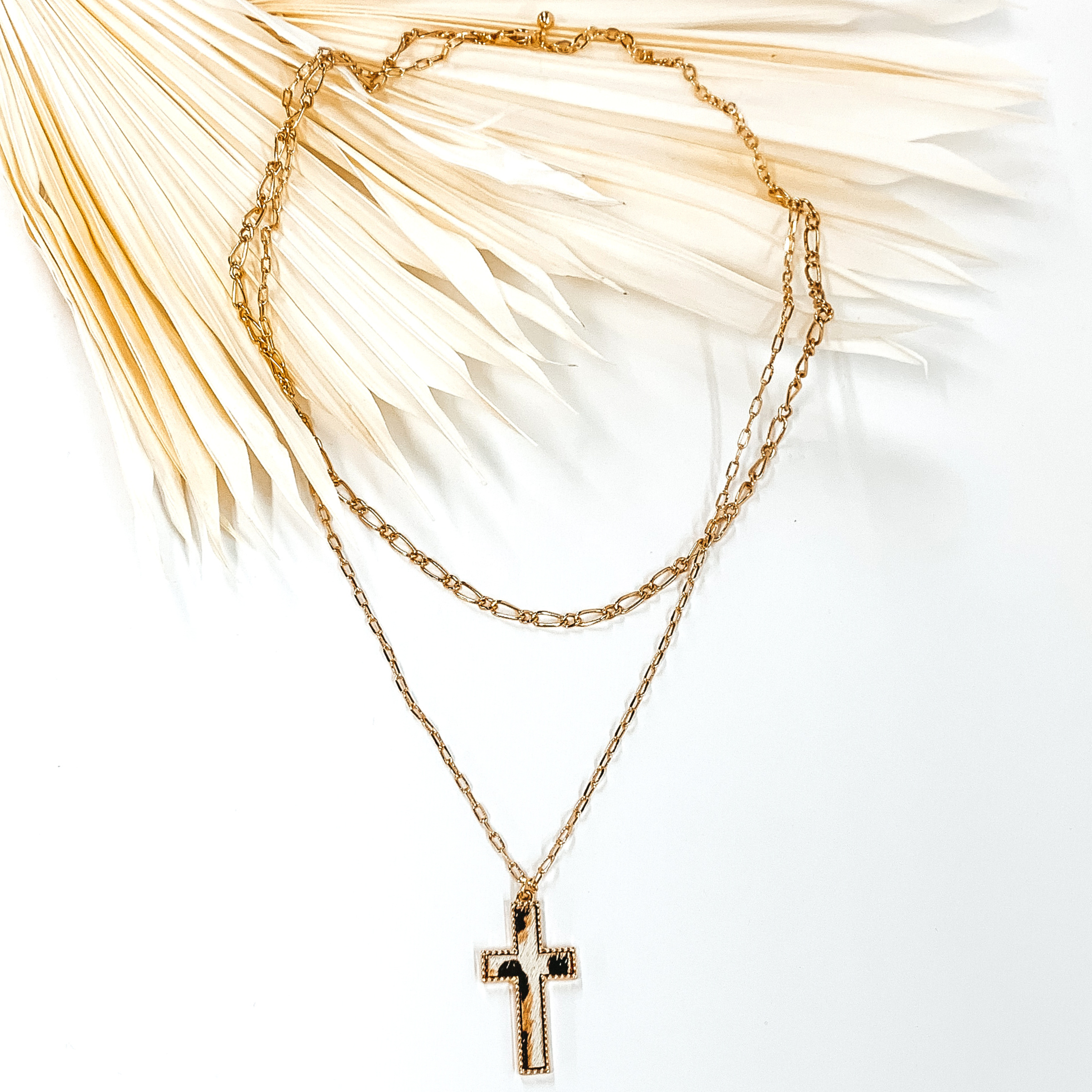 Gold paperclip doubled chain with a cross pendant. The pendant has a white hide inlay with an animal print. This necklace is pictured laying on cream leaf on a white background.