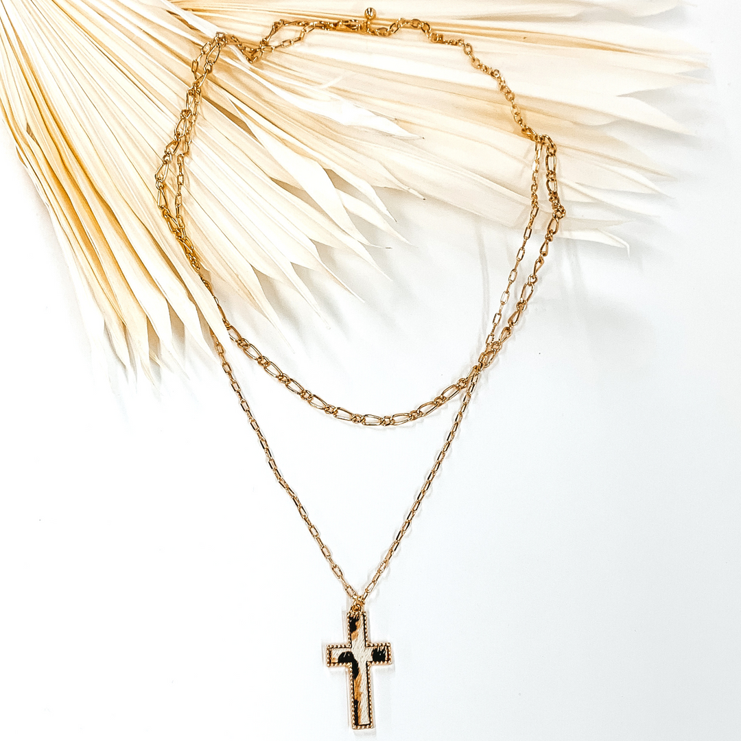 Gold paperclip doubled chain with a cross pendant. The pendant has a white hide inlay with an animal print. This necklace is pictured laying on cream leaf on a white background.
