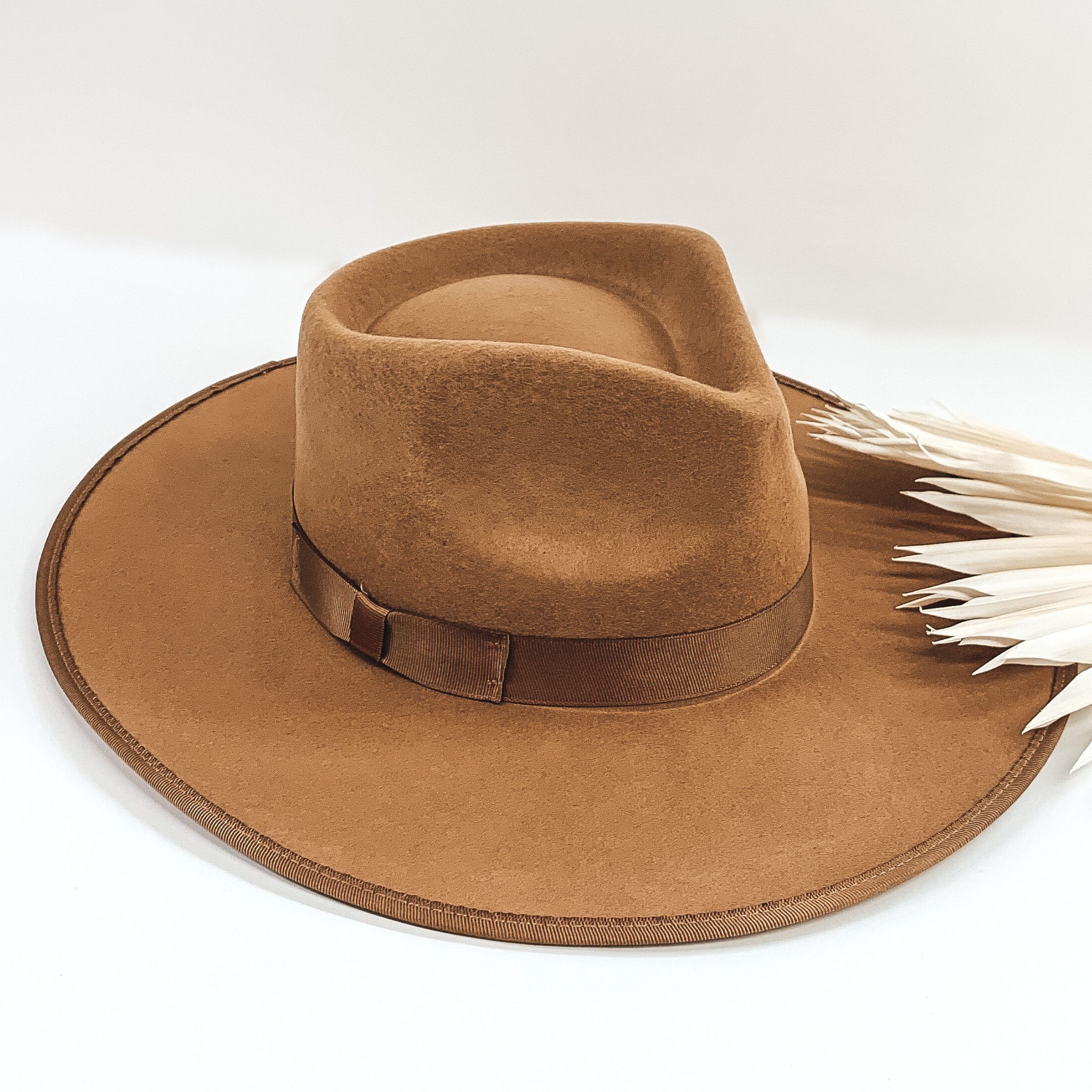 Tan colored hat with a flat brim. The brim has a trimmed outline and the crown has the same colored ribbon wrapped around the bottom. This hat is pictured on a white background with some white leaf decor. 