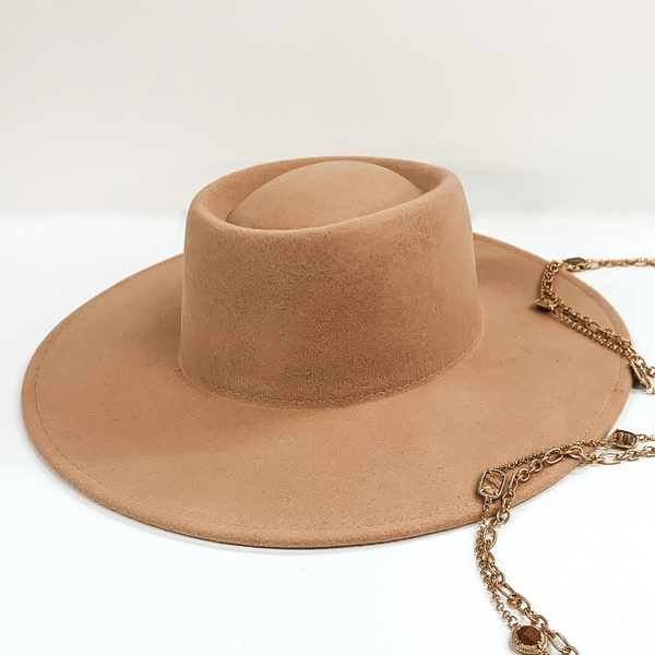 Beige colored hat that has a flat circle bring and an oval crown. This hat is pictured on a white background with a couple gold chained necklaces laying partially on the app. 