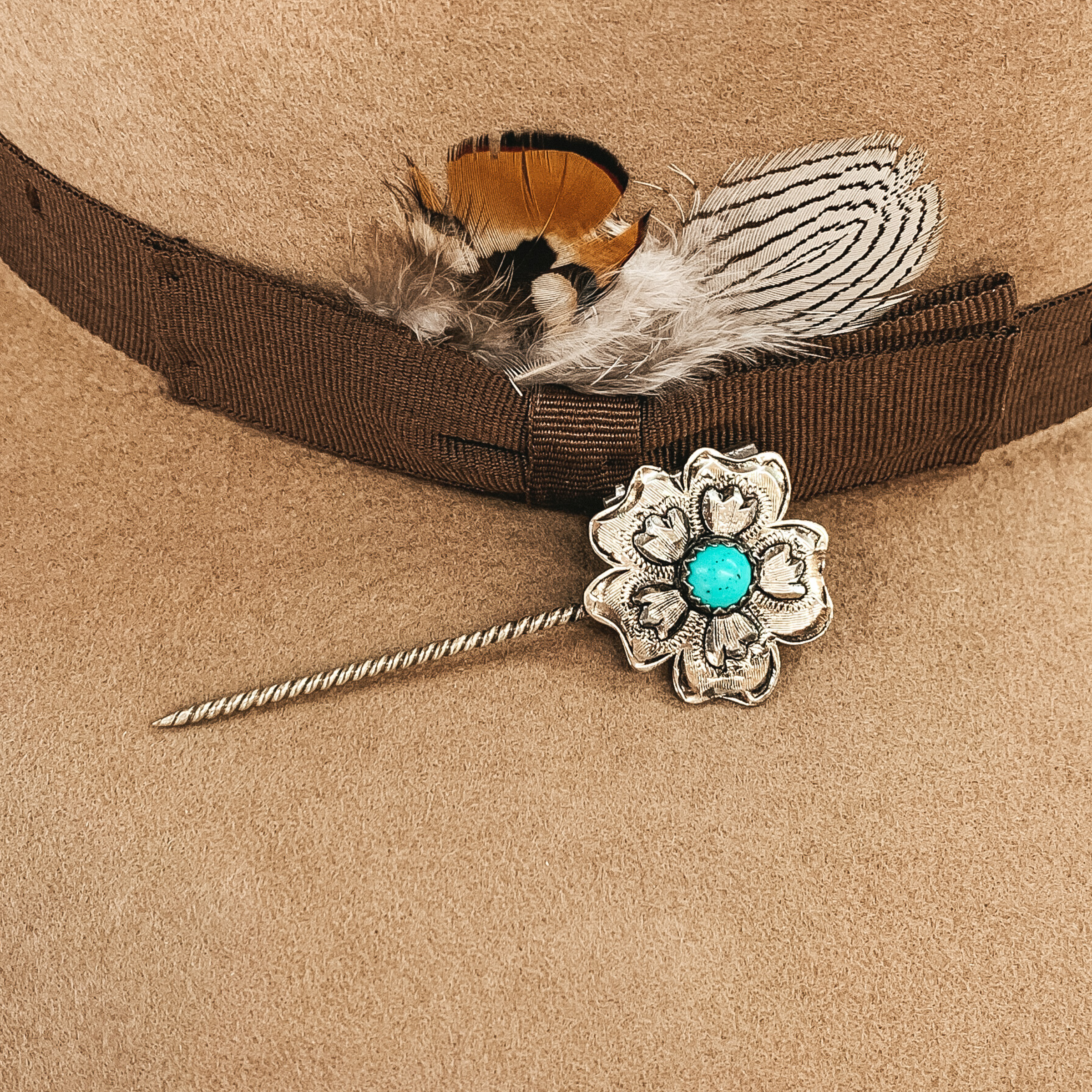 Silver twisted toothpick hat pin with a silver flower pendant. This flower has a lot of small, engraved details around a center turquoise colored stone. This hat pin is pictured on a tan hat.