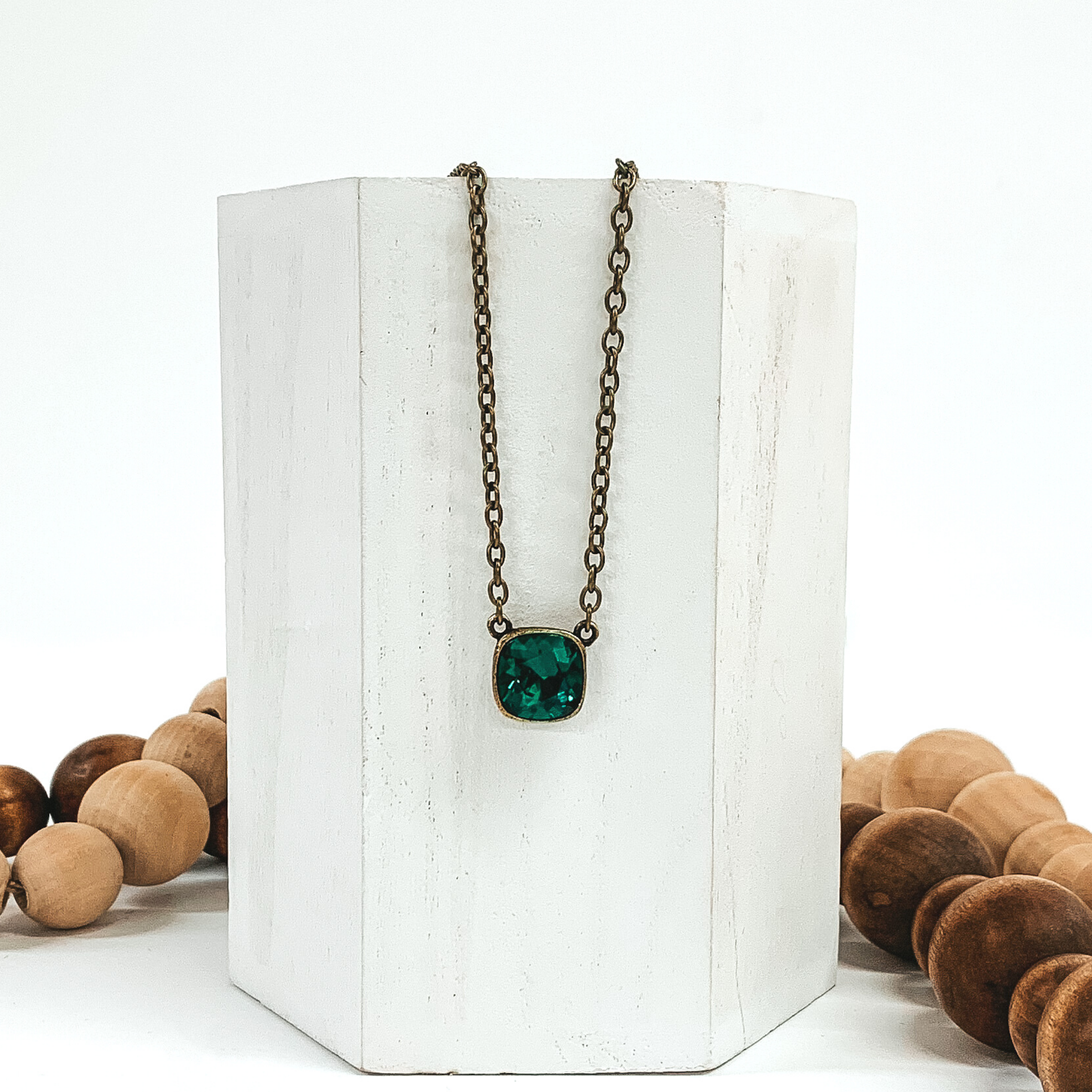 Bronze chained necklace with a square emerald crystal pendant. The necklace is hanging on a white block that is in front of tan and brown beads. 