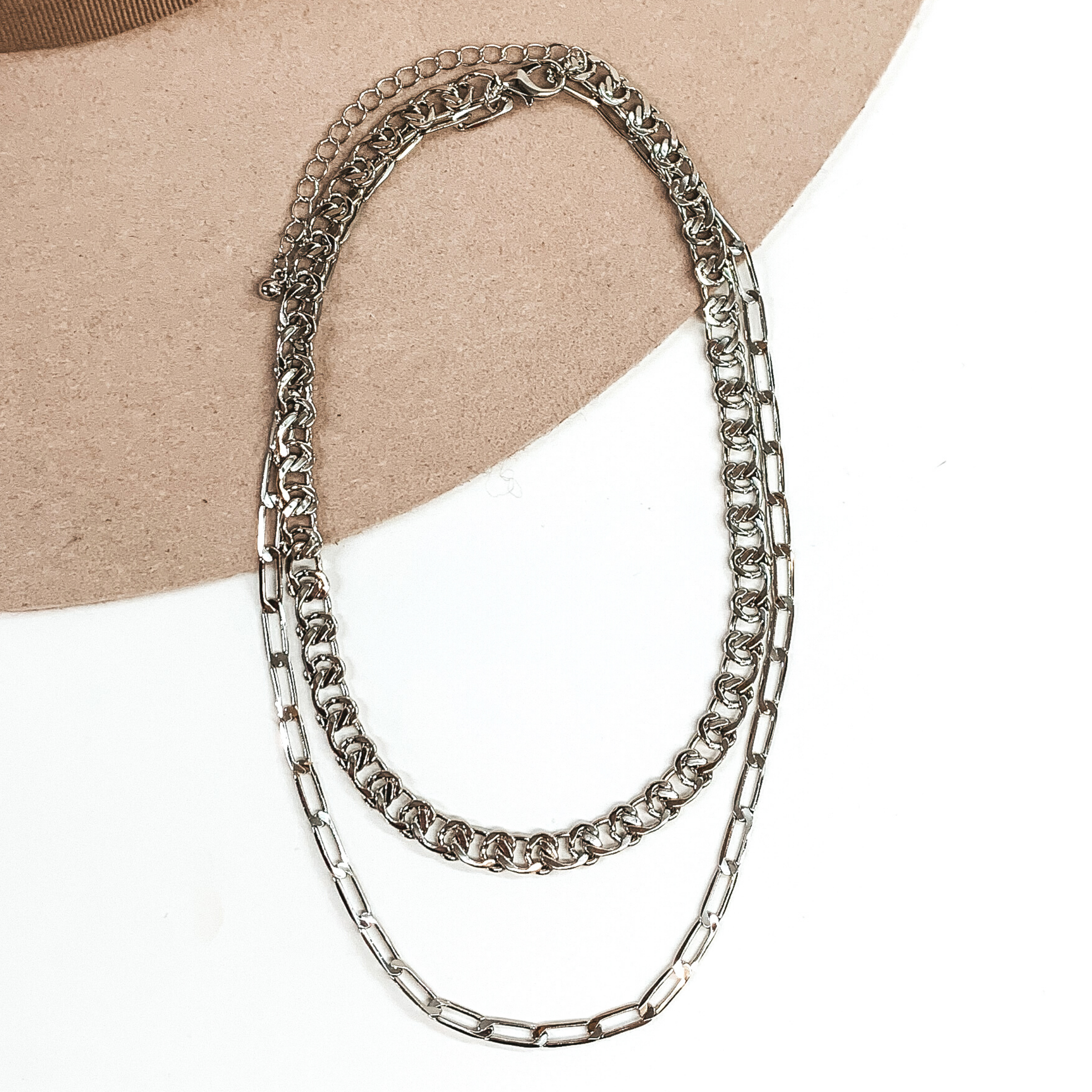 This is a silver double layer, adjustable, chained necklace. There are two different types of chains; a thick curb chain and a thin paperclip chain. this necklace is pictured on a white and beige background. 