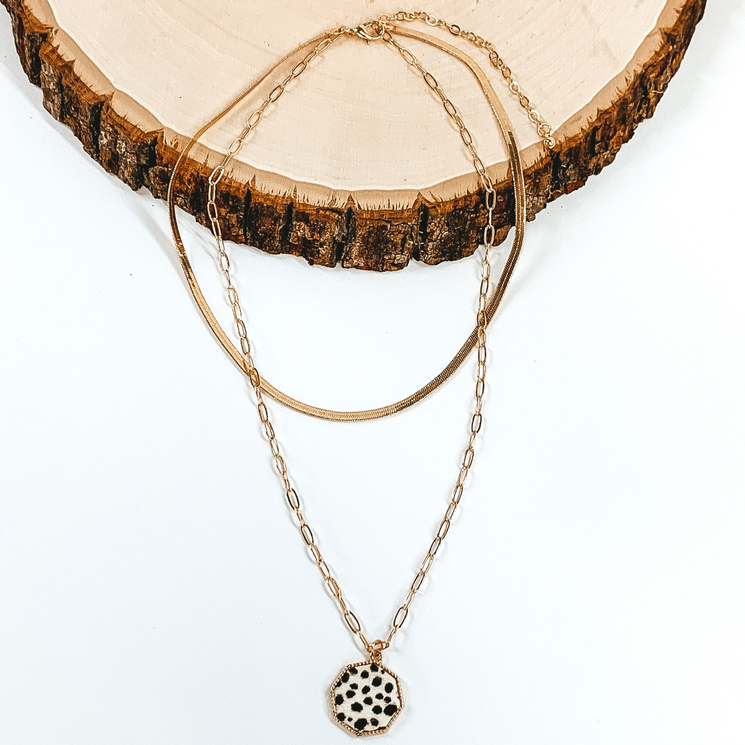Gold paperclip doubled chain with an octogan pendant. The pendant has a white hide inlay with a dotted print. This necklace is pictured laying on a piece of wood on a white background.