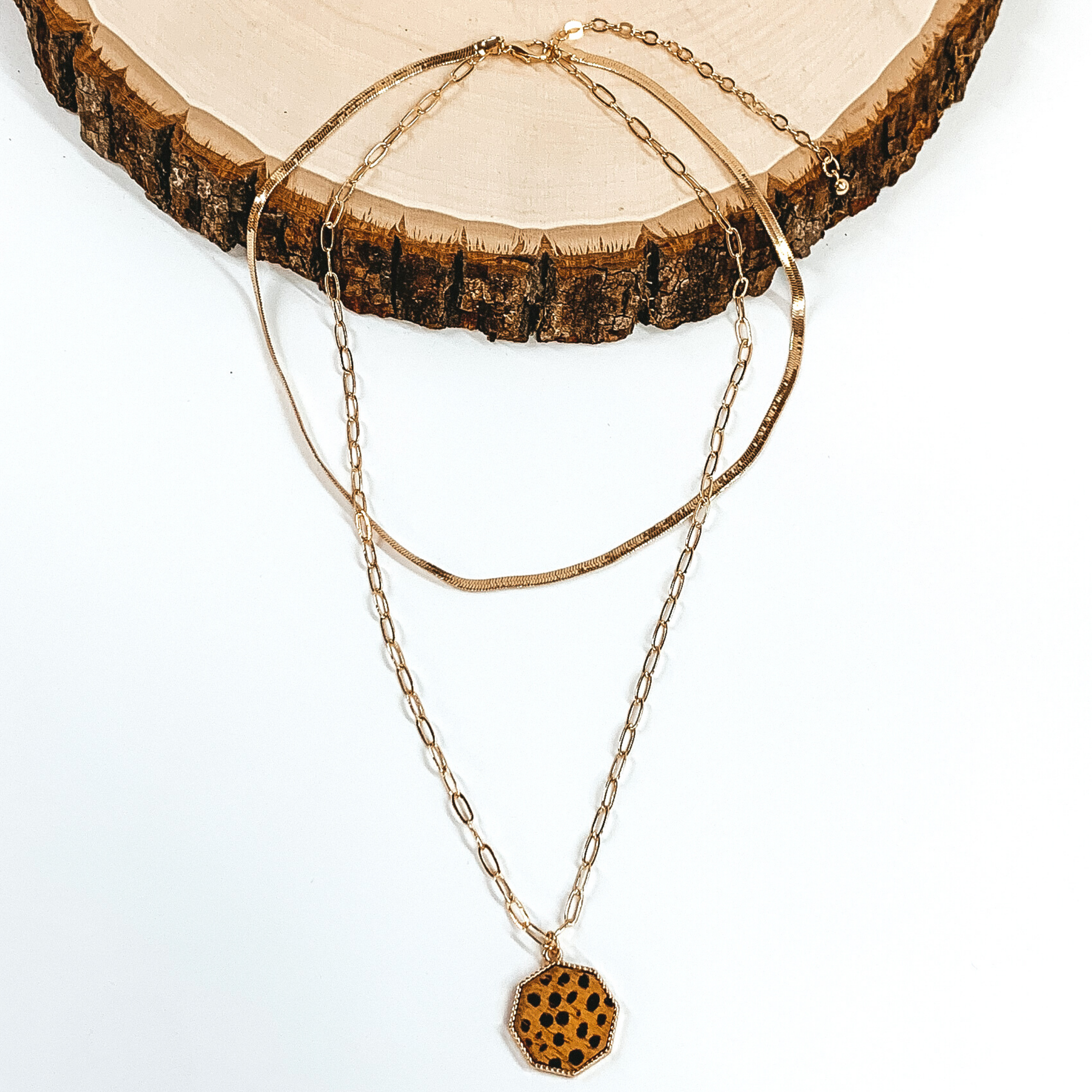 Gold paperclip doubled chain with an octagon pendant. The pendant has a brown hide inlay with a dotted print. This necklace is pictured laying on a piece of wood on a white background.