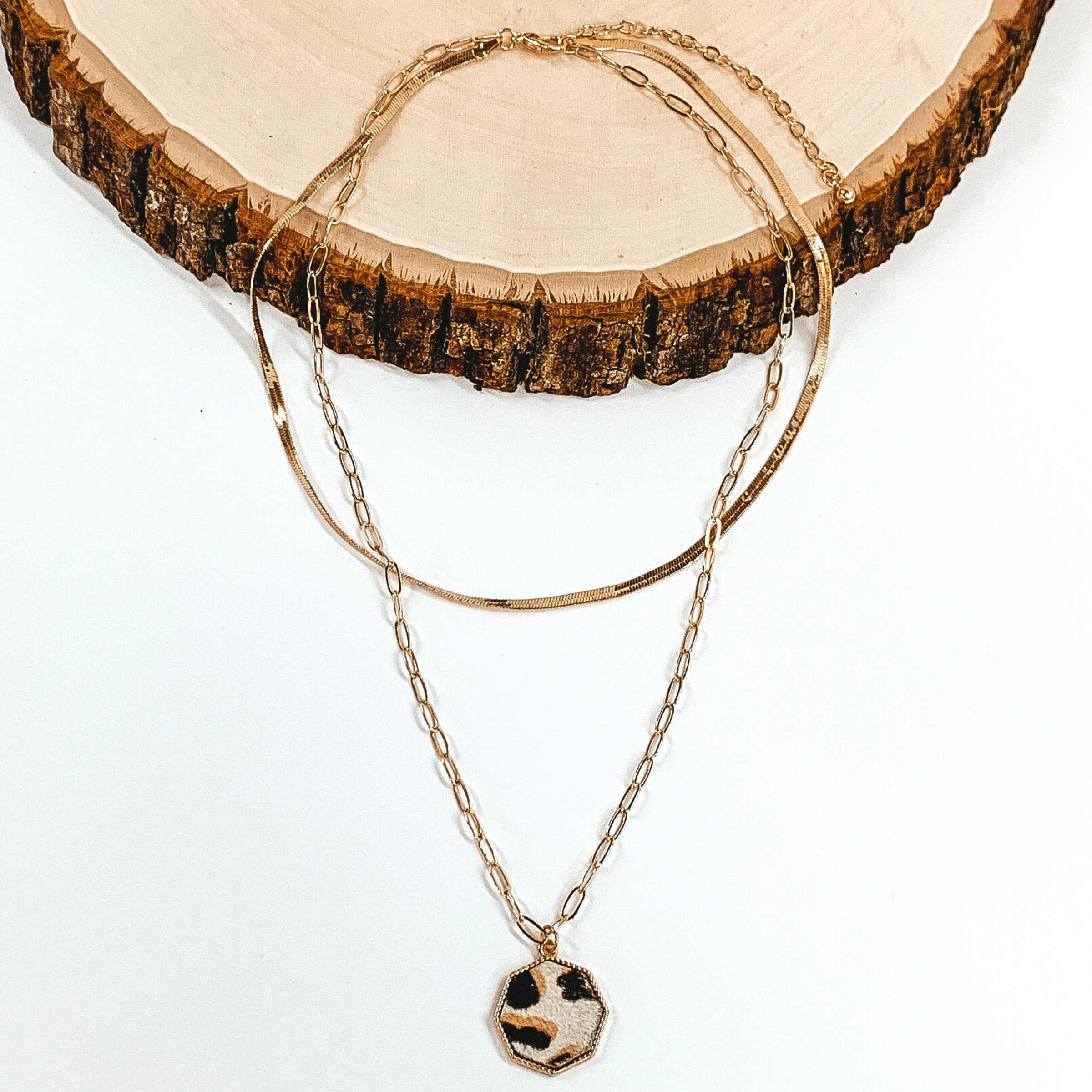 Gold paperclip doubled chain with an octagon pendant. The pendant has a white hide inlay with an animal print. This necklace is pictured laying on a piece of wood on a white background.