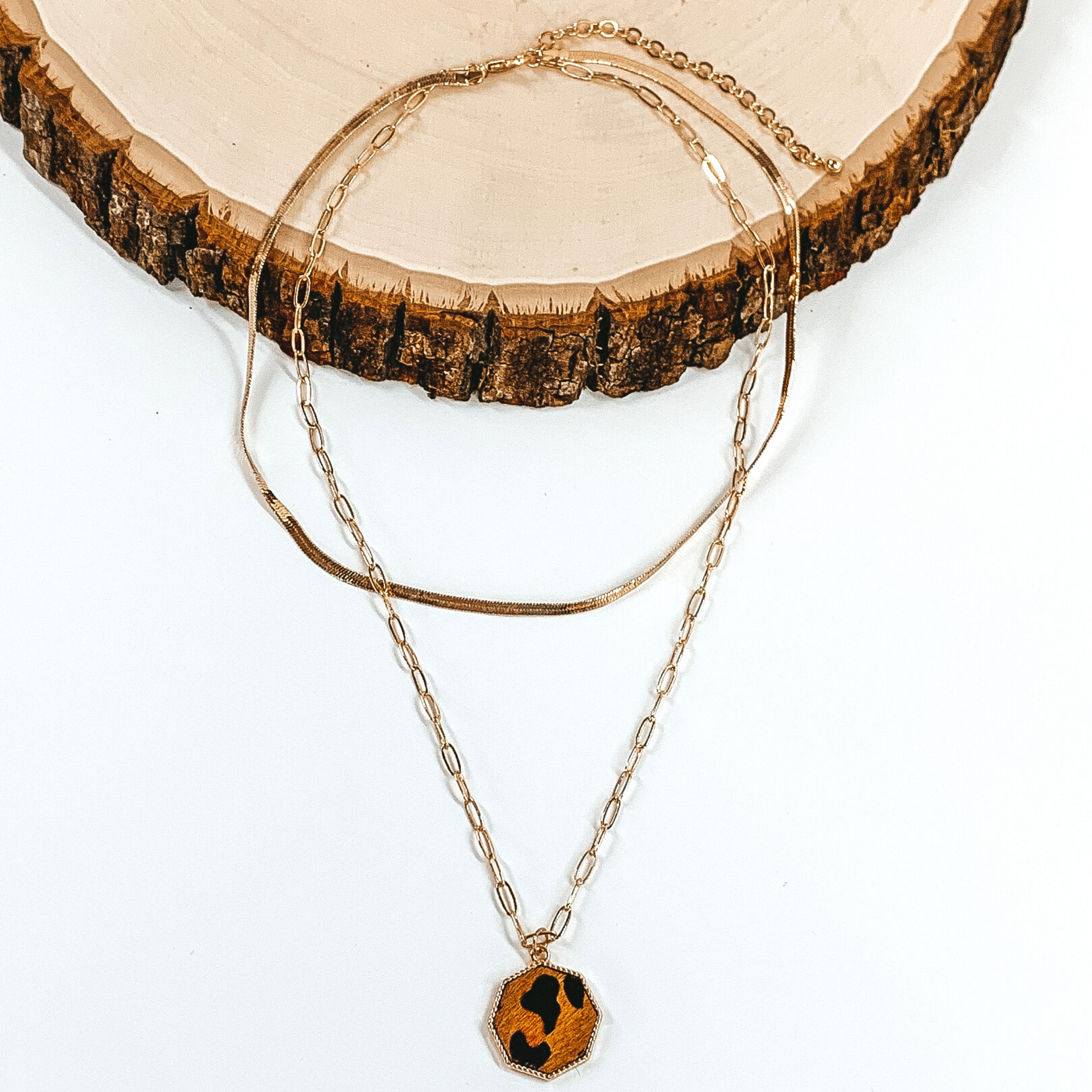 Gold paperclip doubled chain with an octagon pendant. The pendant has a brown hide inlay with an animal print. This necklace is pictured laying on a piece of wood on a white background.