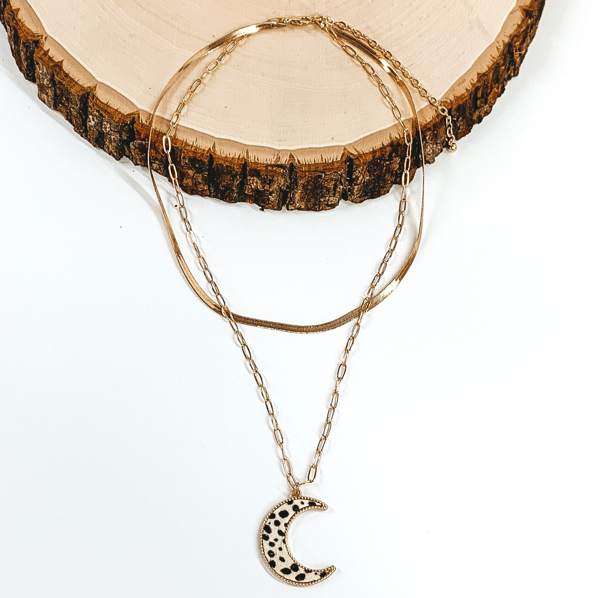 Gold paperclip doubled chain with an moon pendant. The pendant has a white hide inlay with a dotted print. This necklace is pictured laying on a piece of wood on a white background.
