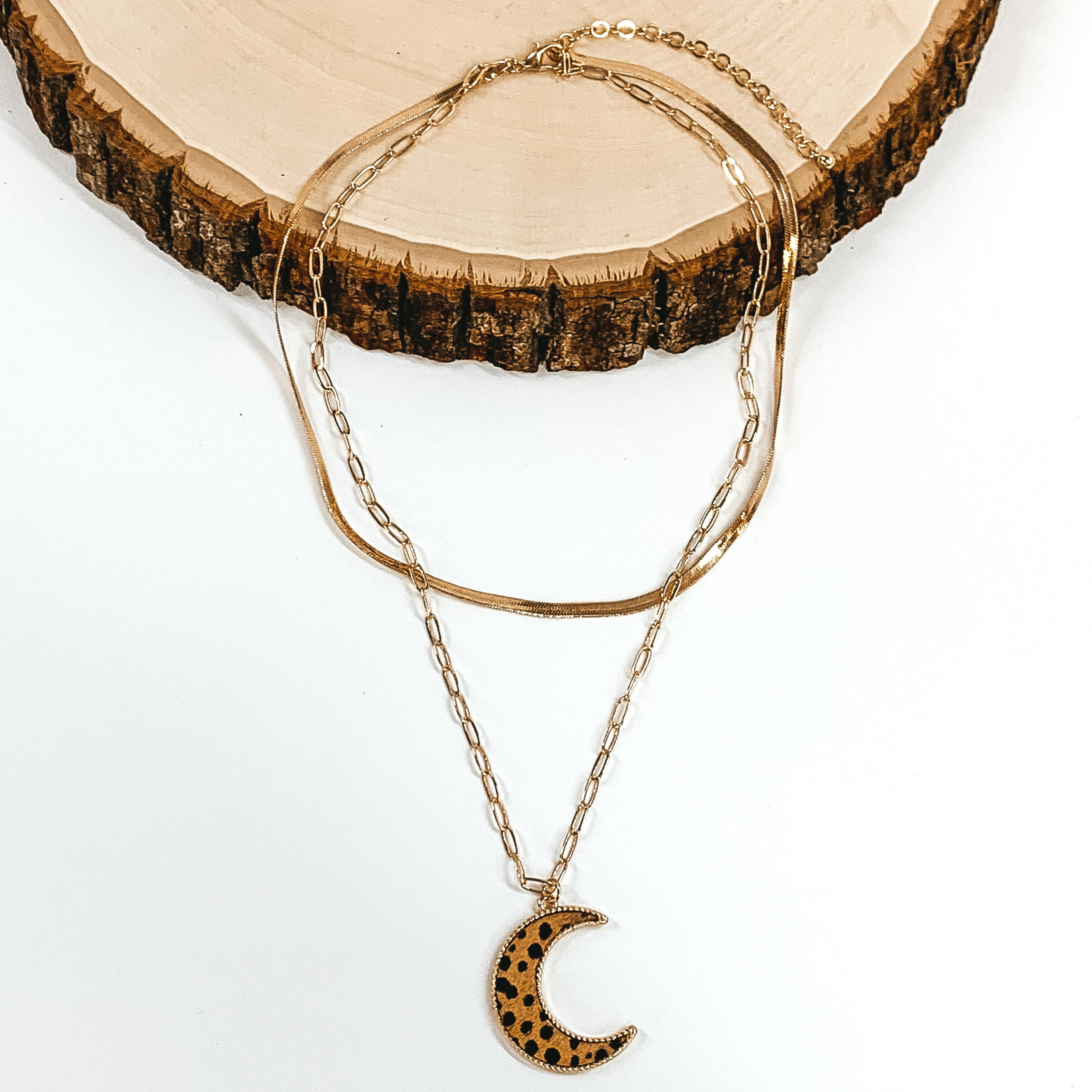 Gold paperclip doubled chain with an moon pendant. The pendant has a brown hide inlay with a dotted print. This necklace is pictured laying on a piece of wood on a white background.