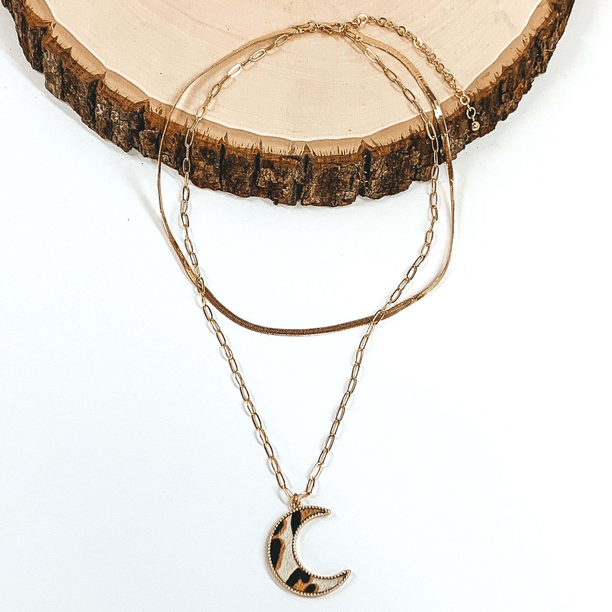 Gold paperclip doubled chain with a moon pendant. The pendant has a white hide inlay with an animal print. This necklace is pictured laying on a piece of wood on a white background.