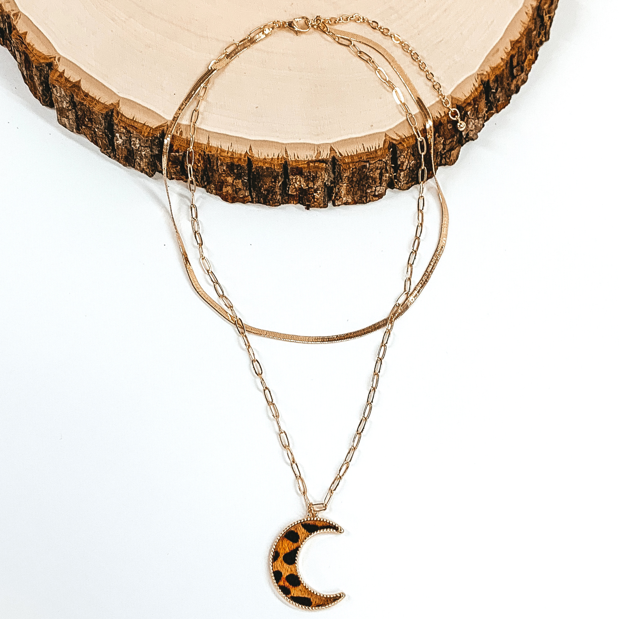 Gold paperclip doubled chain with a moon pendant. The pendant has a brown hide inlay with an animal print. This necklace is pictured laying on a piece of wood on a white background.