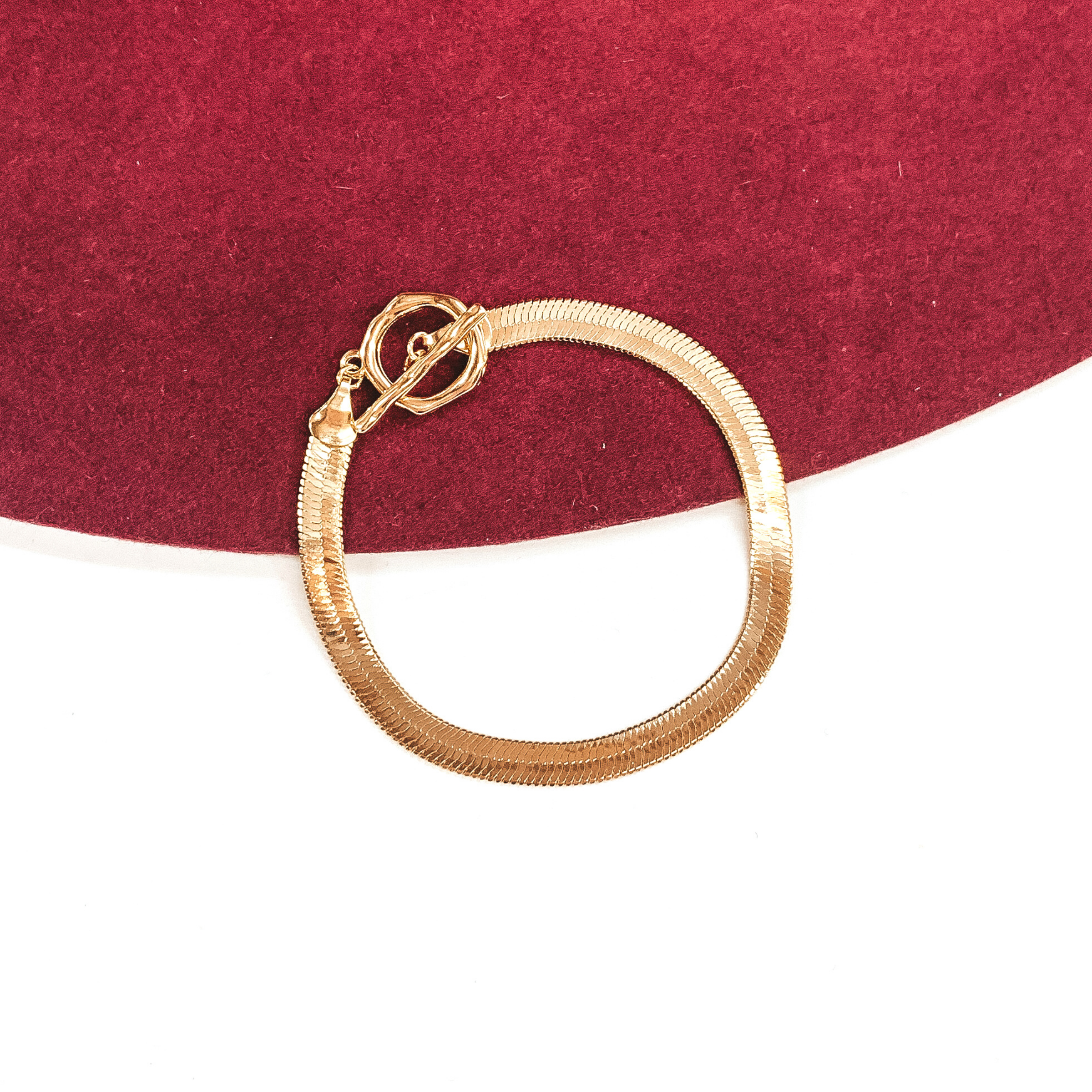Gold herringbone bracelet that closes with a toggle clasp. This bracelet is pictured on a white and red background.