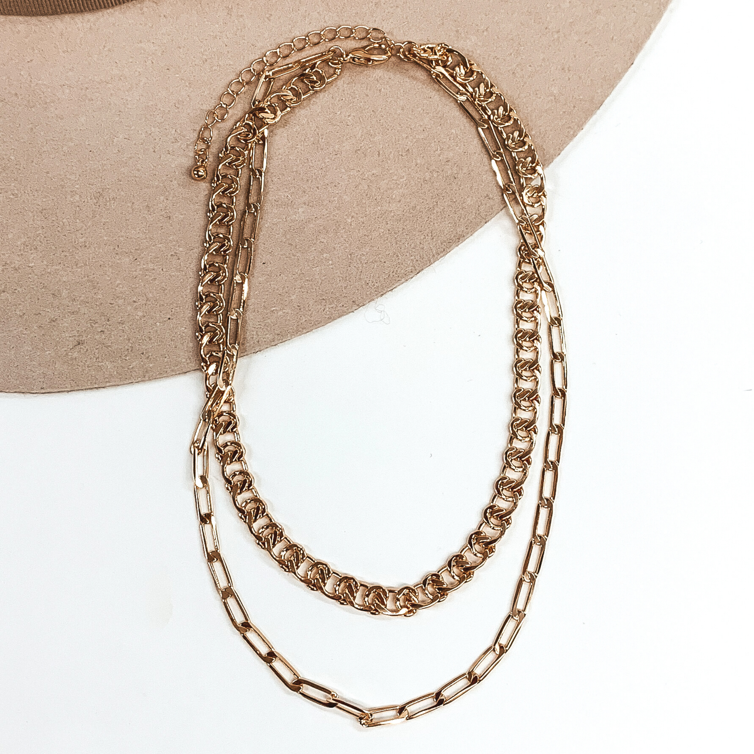 This is a gold double layer, adjustable, chained necklace. There are two different types of chains; a thick curb chain and a thin paperclip chain. this necklace is pictured on a white and beige background.