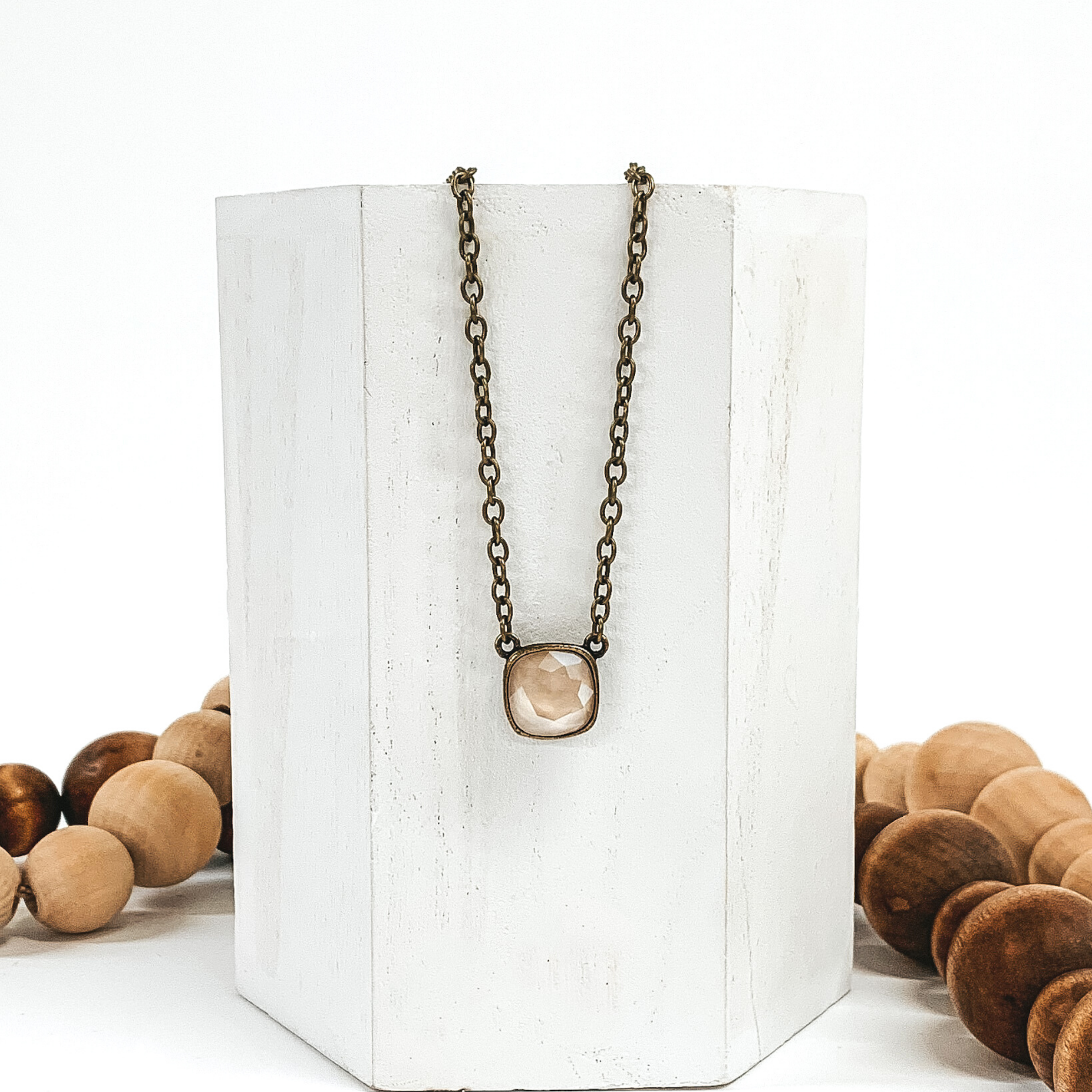 Bronze chained necklace with a square ivory crystal. This necklace is hanging on a white block that is in front of brown and tan beads. 