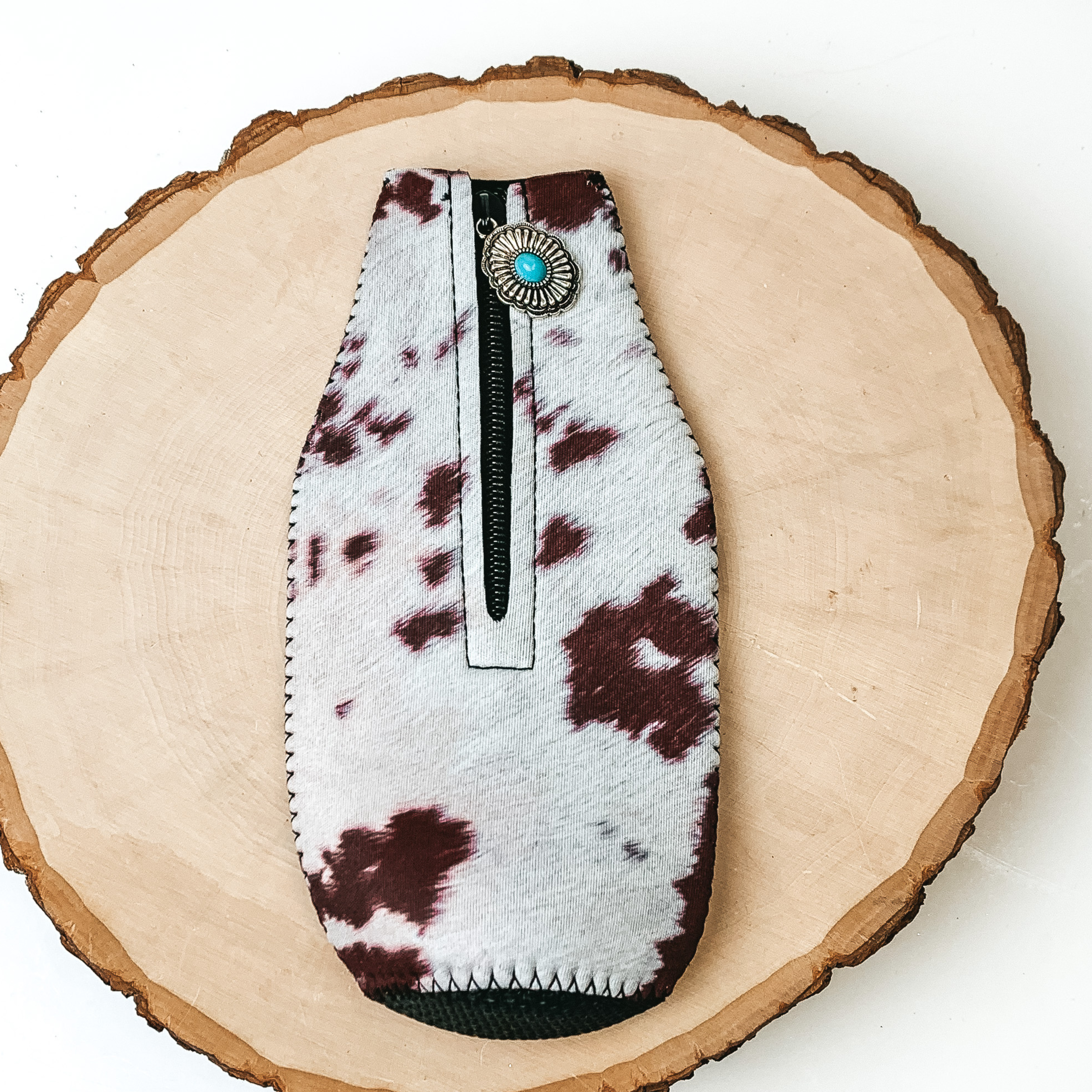 This Cowhide Print Zip Up kooozie With a Concho Charm is pictured on a peice of wood, with a white background.  
