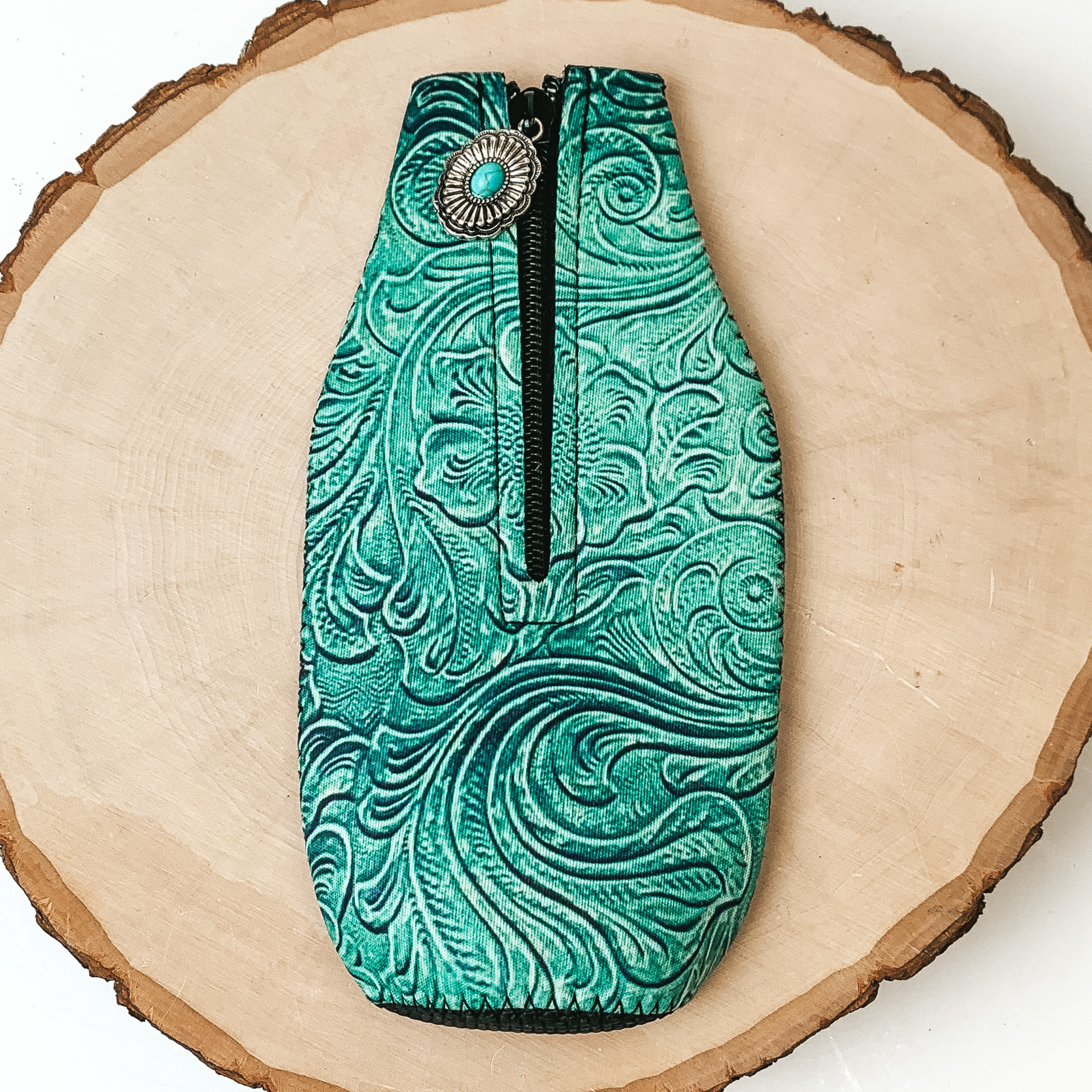 This Turquoise Leather Tooled Print Zip Up kooozie With a Concho Charm is pictured on a peice of wood, with a white background.  