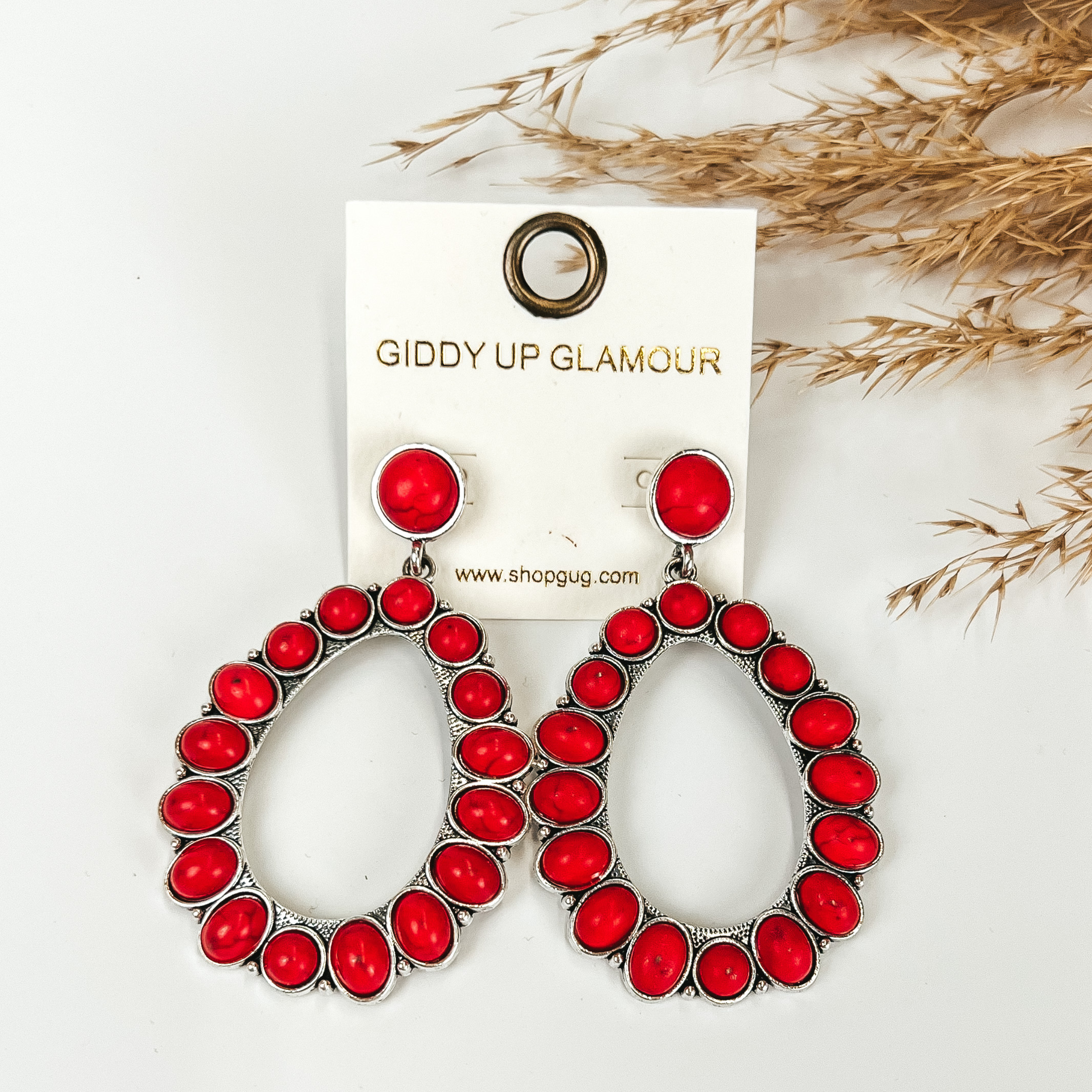 Red stoned teardrop earrings, with pompous grass in the top right hand corner, on a white background. 