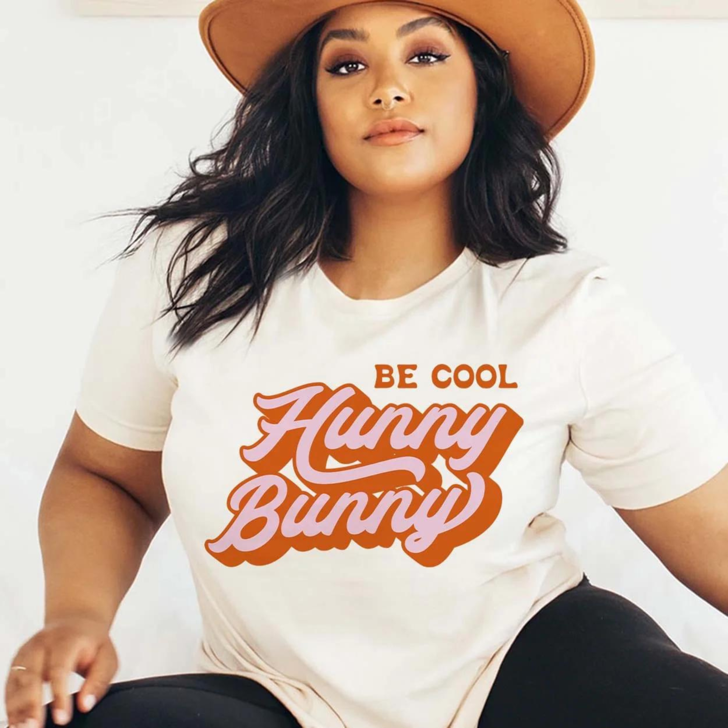 A cream colored short sleeve tee with the words "Be cool hunny bunny". Be cool is in a standard font in orange while the rest is pink with an orange shadow in cursive. Item is pictured on a plain white background.