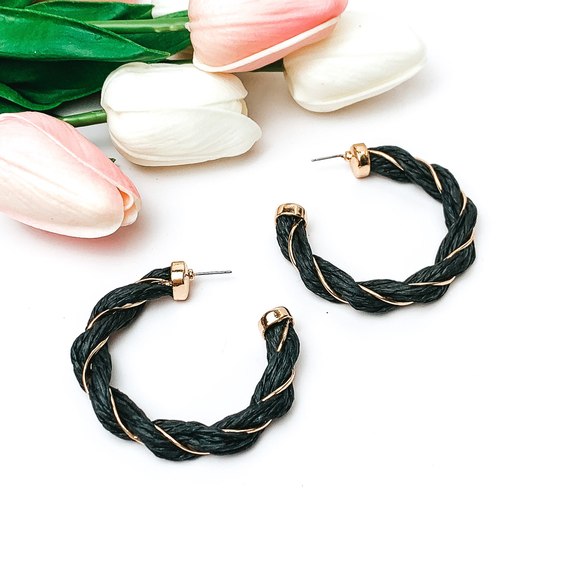 Pictured are black raffia twisted hoop earrings with gold detailing.  They are pictured with pink and white tulips on a white background.