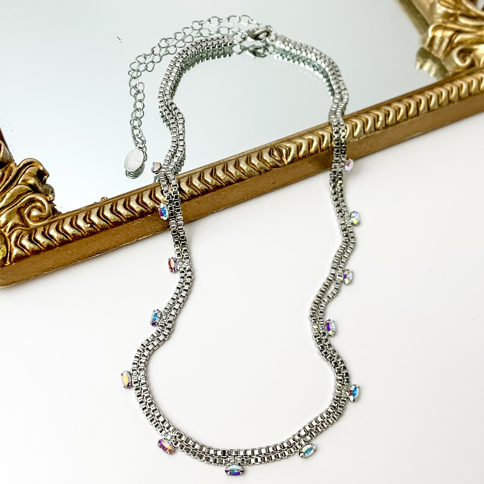 Pictured is a double, silver box chain necklace with ab crystal charms. This necklace is pictured partially laying on a gold mirror on a white background.    