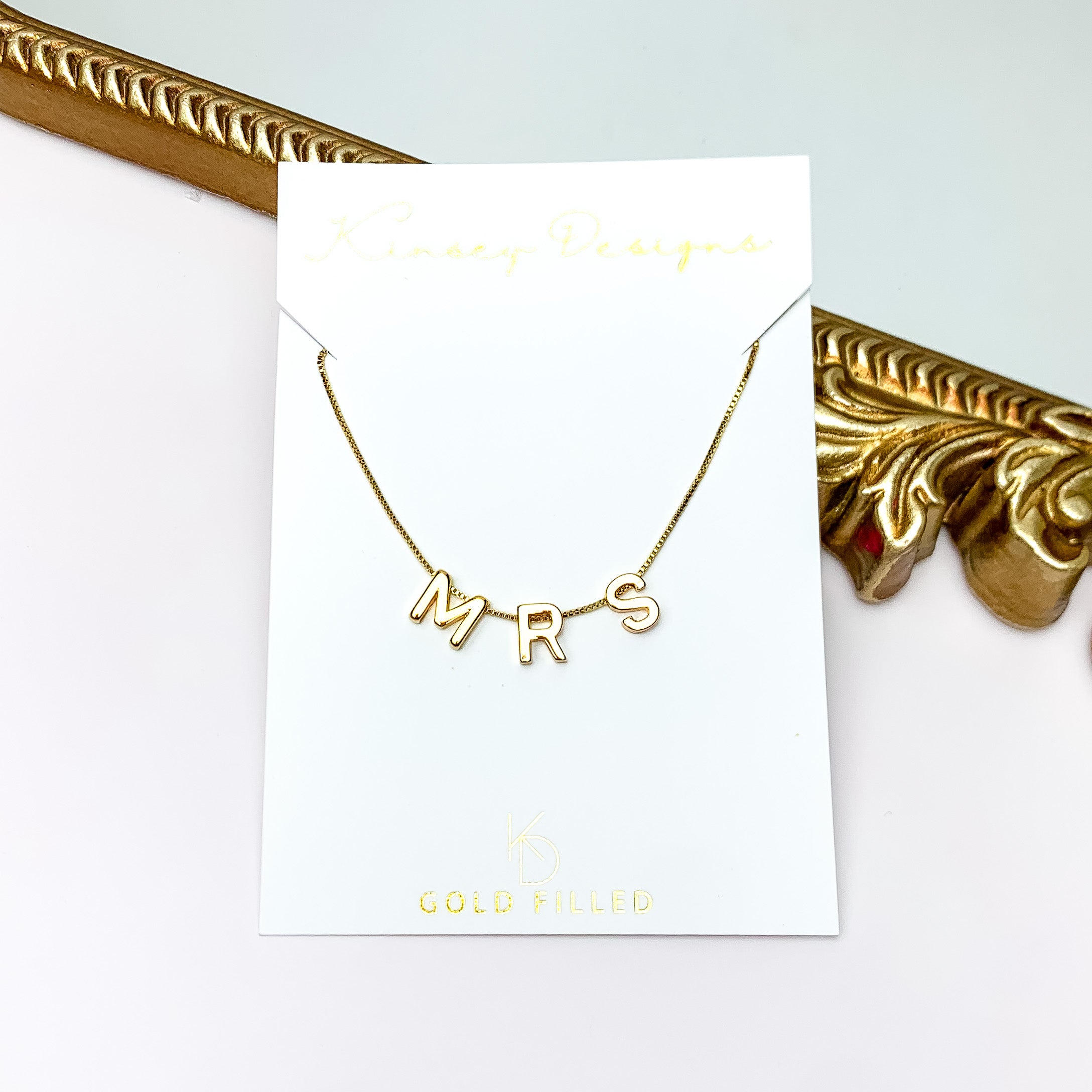 Pictured is a gold chain necklace with gold letter charms. This necklace includes letters that spell out "MRS". This necklace is pictured on a white necklace holder that is in front of a gold mirror on a white background.  