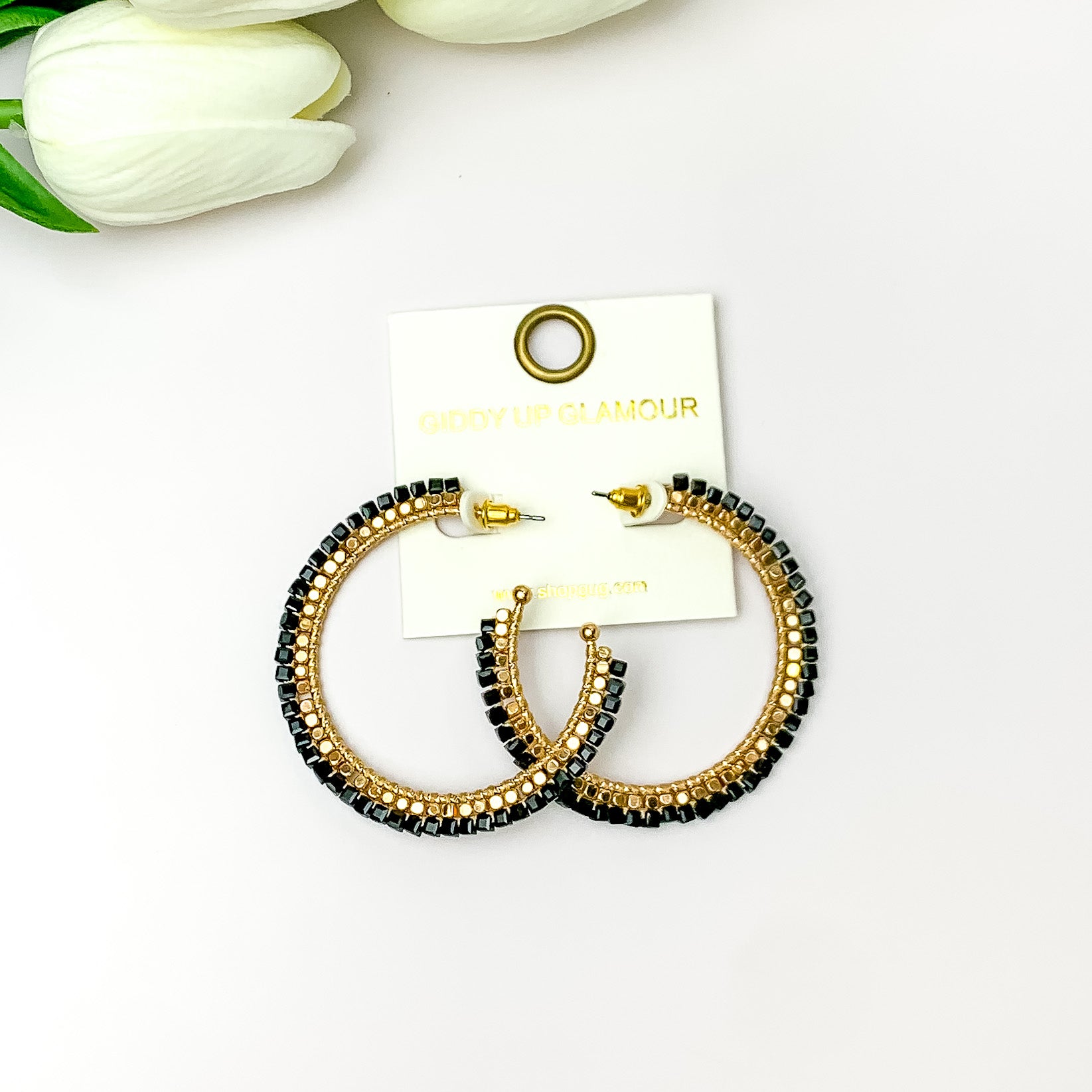 Pictured are circle gold toned hoop earrings with gold beads around it and black crystal outline. They are pictured with white flowers on a white background.